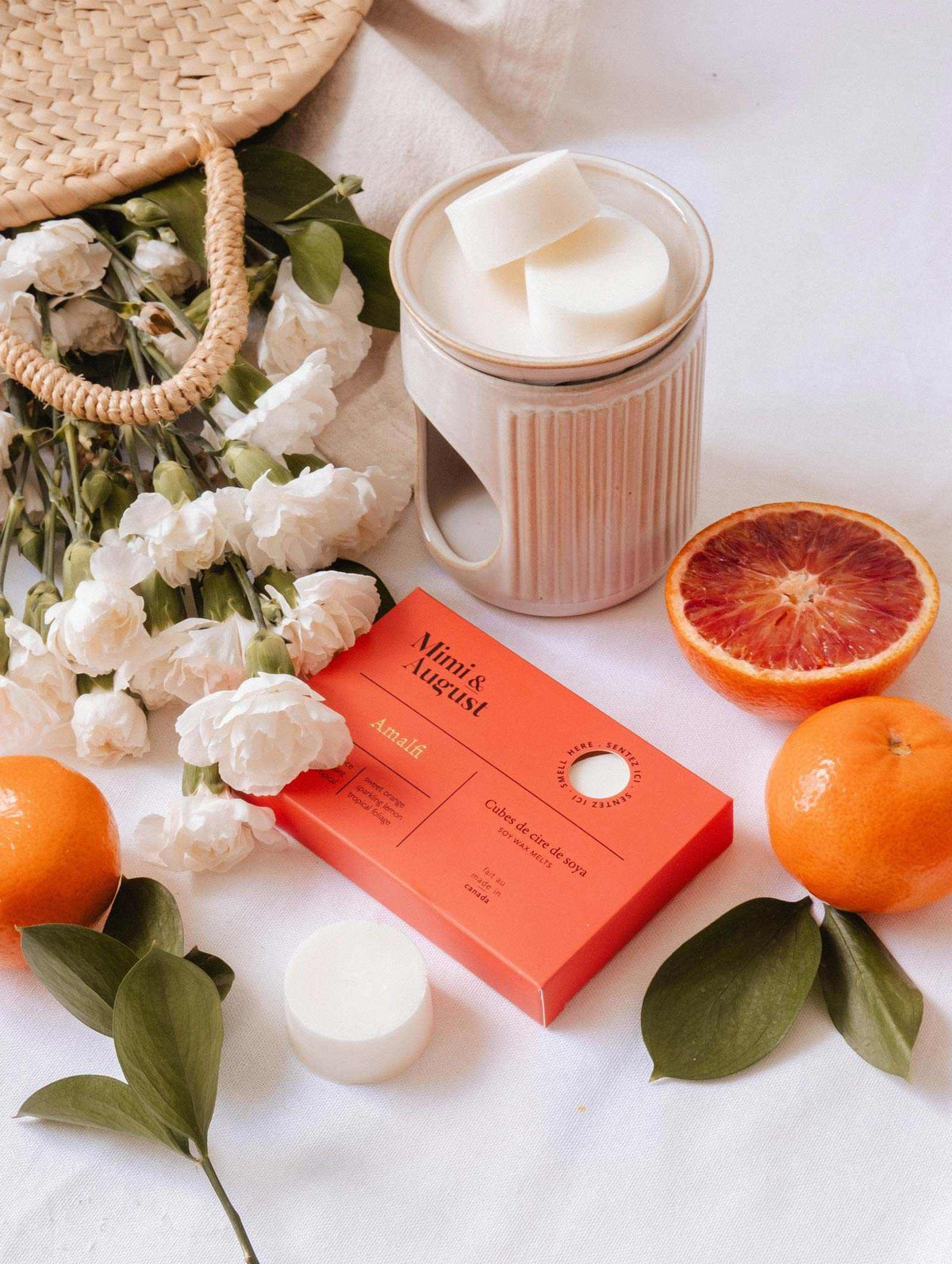 A ceramic mug, red soap box, white flowers, woven bag, orange, tangerine, and sliced blood orange on a white surface reflect the lasting scent of Mimi & August’s Soy Wax Melts - Amalfi made with natural soy wax.