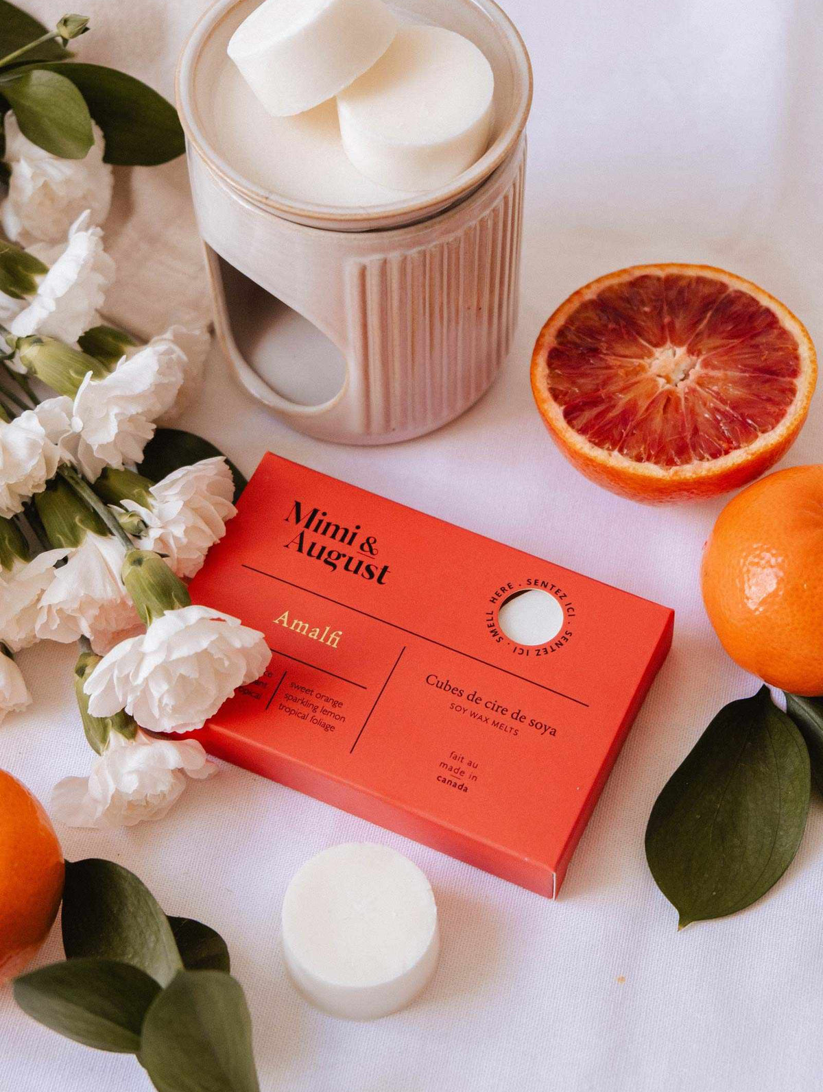 The Mimi & August Soy Wax Melts - Amalfi set offers a long-lasting fragrance and comes with a red gift box, ceramic wax warmer, white flowers, green leaves, an orange, and a halved blood orange on a white surface.