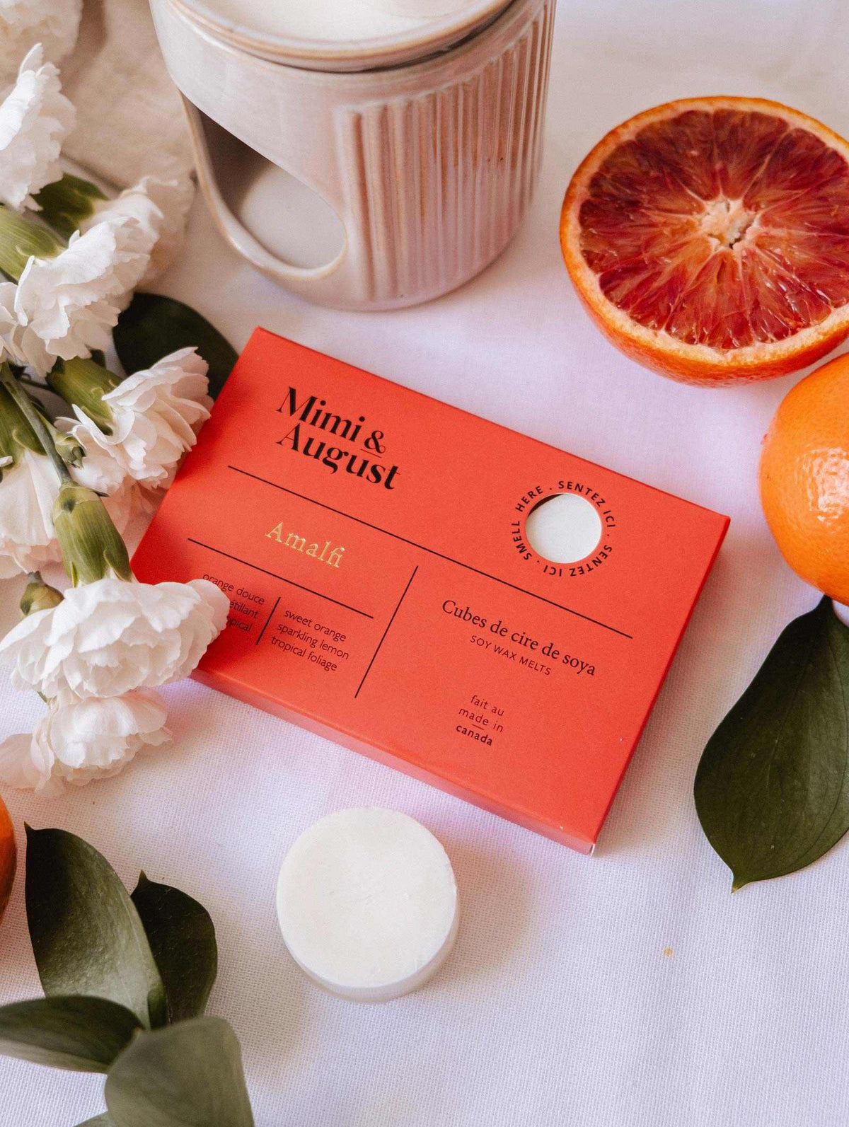 A box of Mimi & August Soy Wax Melts - Amalfi sits on a table with white flowers, orange slices, green leaves, and a ceramic wax warmer, offering a long-lasting natural fragrance from soy wax.