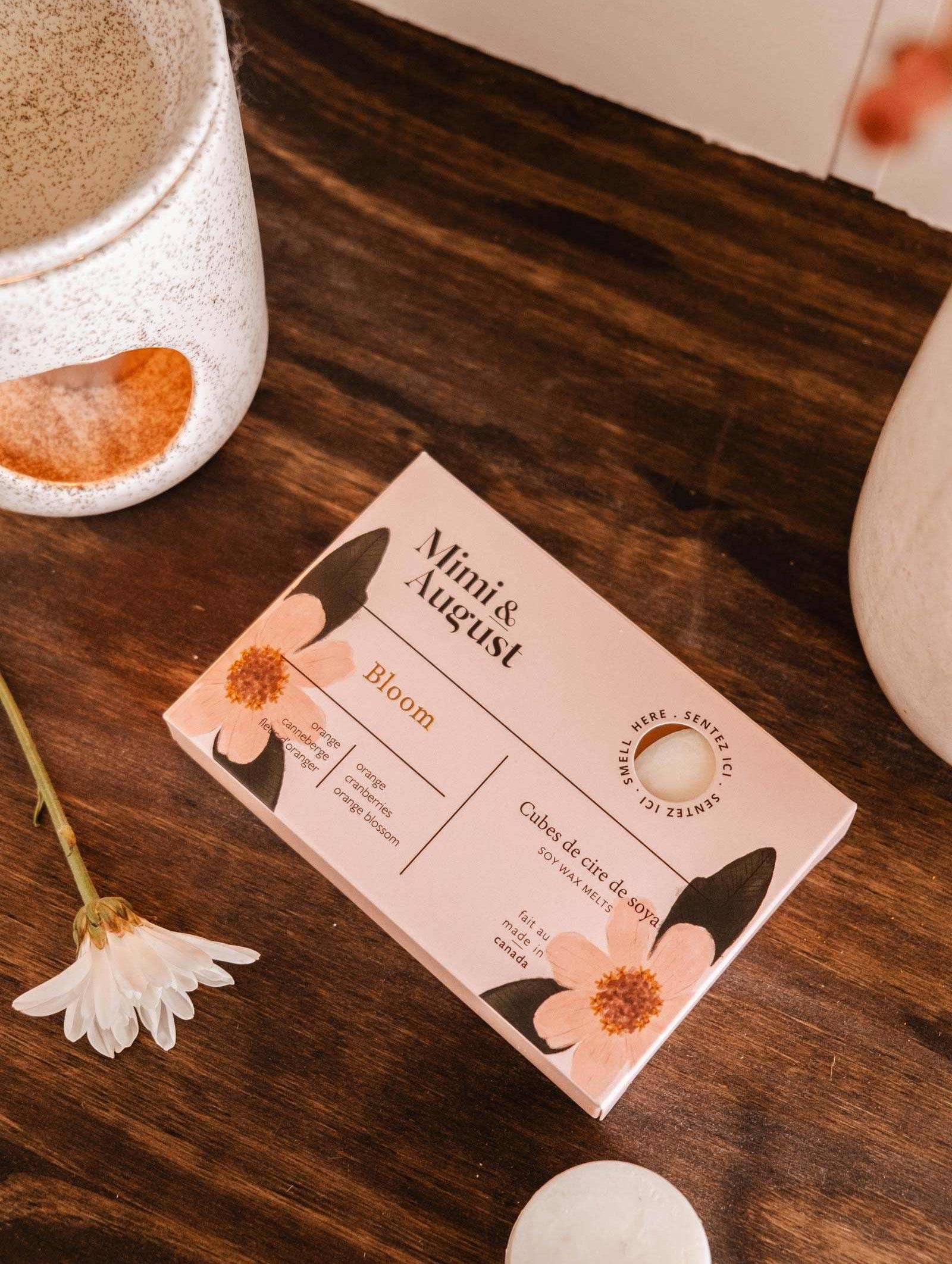 A box of "Mimi & August" Soy Wax Melts - Bloom sits on a wooden surface, surrounded by ceramic cups and a small white flower, exuding an eco-friendly charm akin to a solid face cleanser.