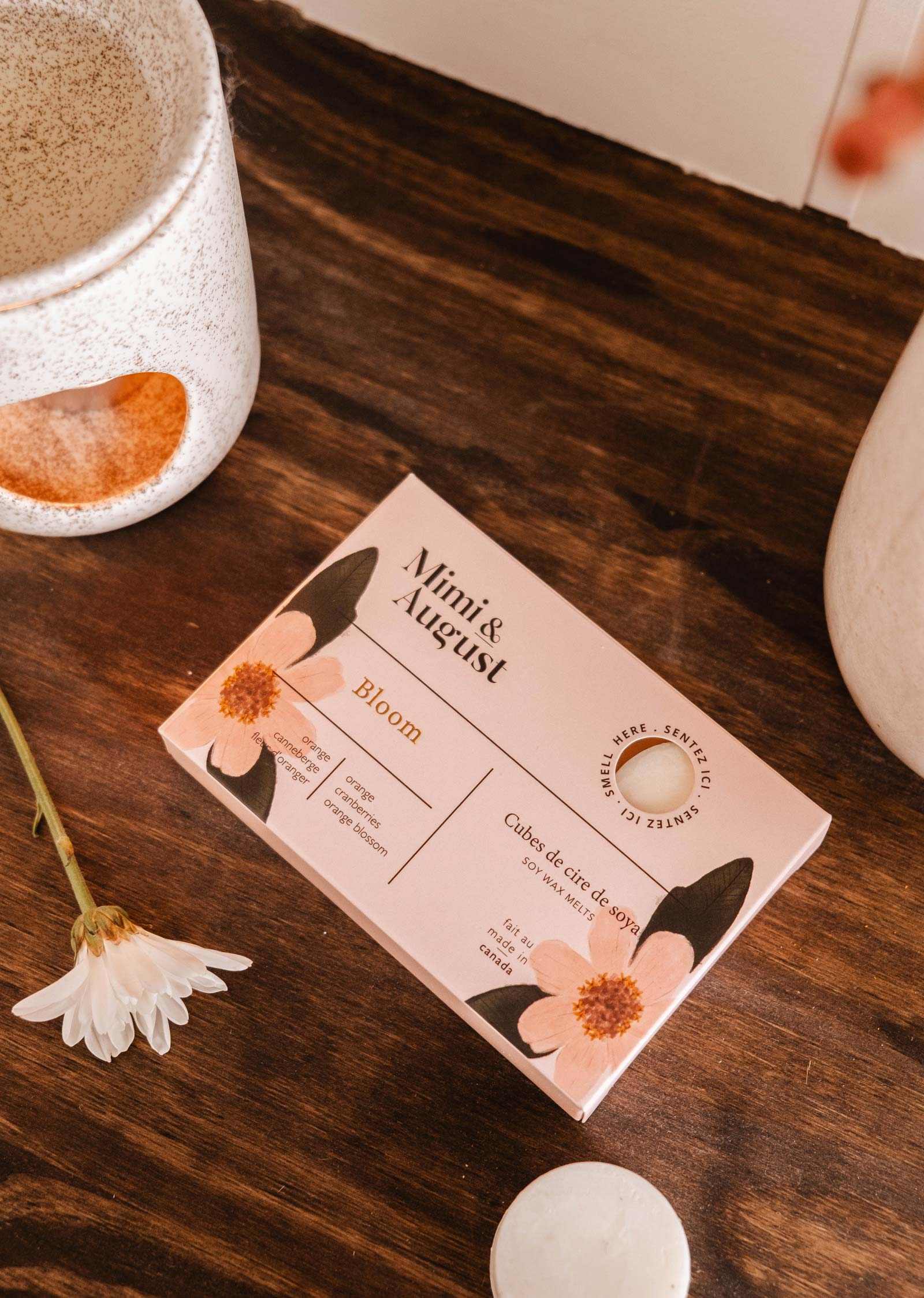Une boîte de fondants de cire de soya Bloom de Mimi & August repose sur une surface en bois, entourée de tasses en céramique et d'une petite fleur blanche, dégageant un charme écoresponsable.