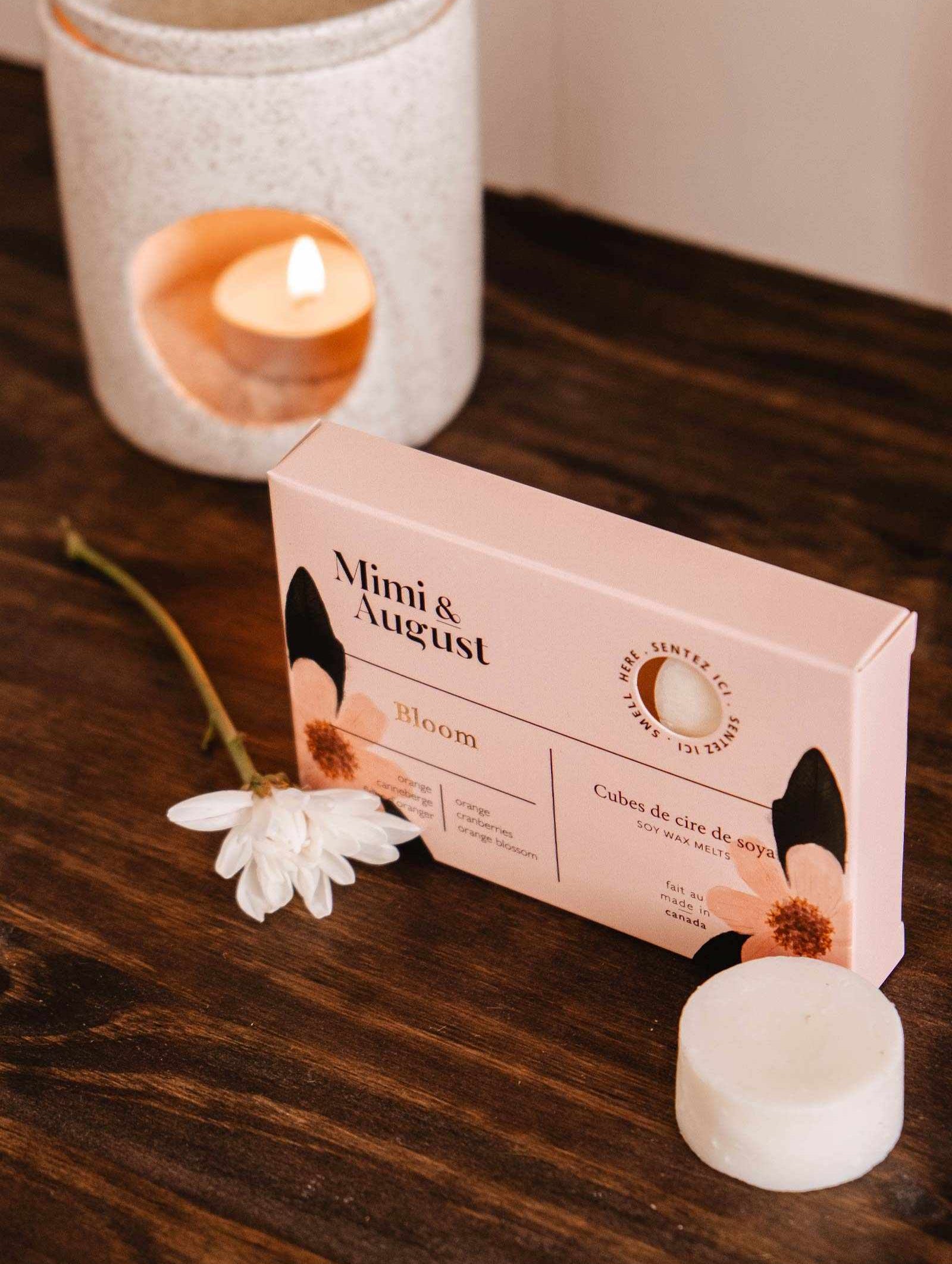 A "Mimi & August Bloom" box sits on a wooden surface with a white flower, while an eco-friendly soy wax melt and candle holder with a lit candle offer long-lasting fragrance in the background.