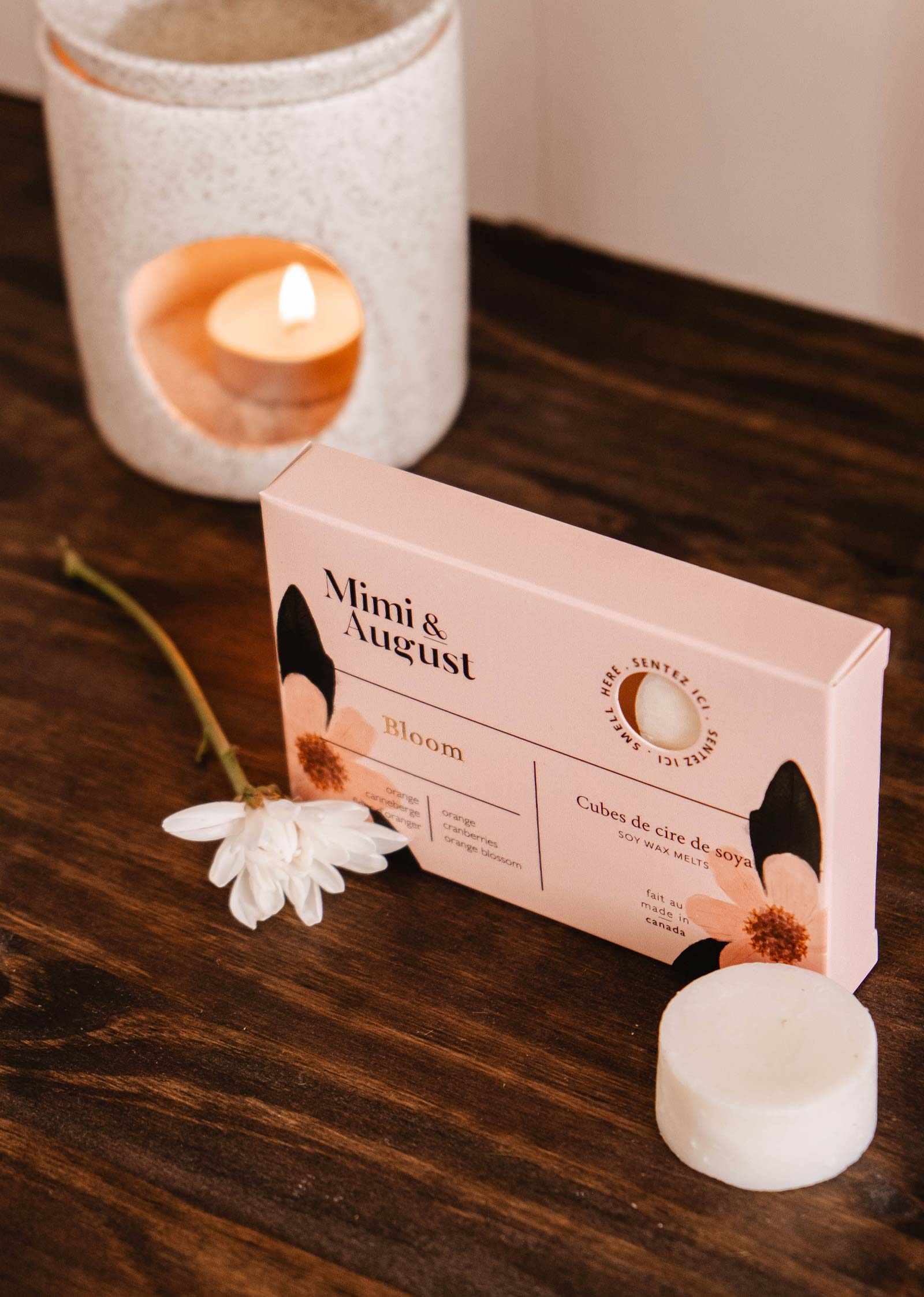 Une boîte « Mimi & August Bloom » repose sur une surface en bois avec une fleur blanche, tandis qu'un fondant de cire de soya écoresponsable et un bougeoir avec une bougie allumée offrent un parfum longue durée en arrière-plan.
