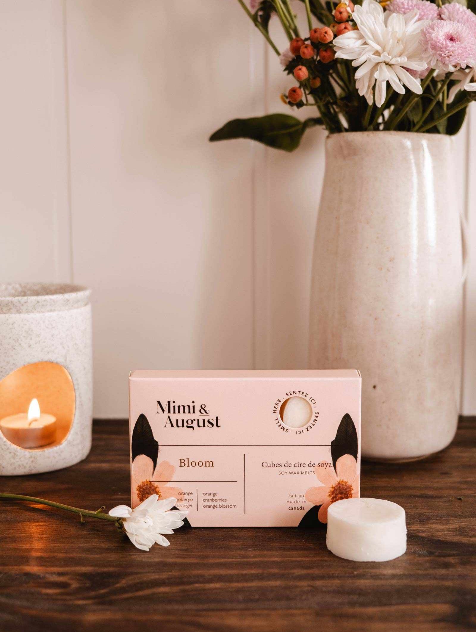 On a wooden table, surrounded by flowers, a small white candle, and a vase of mixed blooms, sits a box of Mimi & August Soy Wax Melts - Bloom. These eco-friendly cubes offer a long-lasting fragrance for any room.
