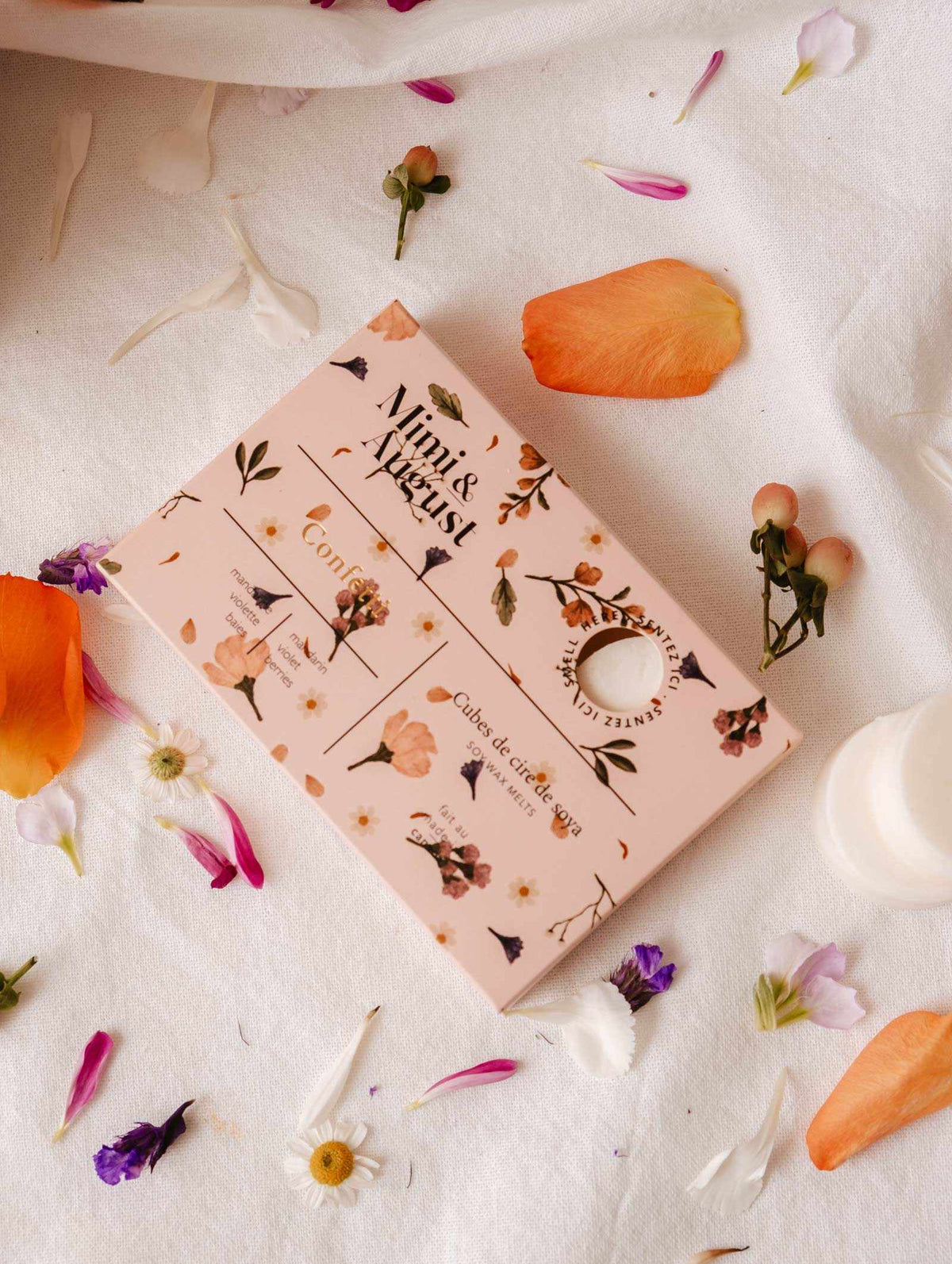 A pink box with floral patterns labeled "Mimi & August" rests on white fabric surrounded by colorful petals and small flowers, offering a long-lasting fragrance with an eco-friendly touch. Product: Soy Wax Melts - Confetti.