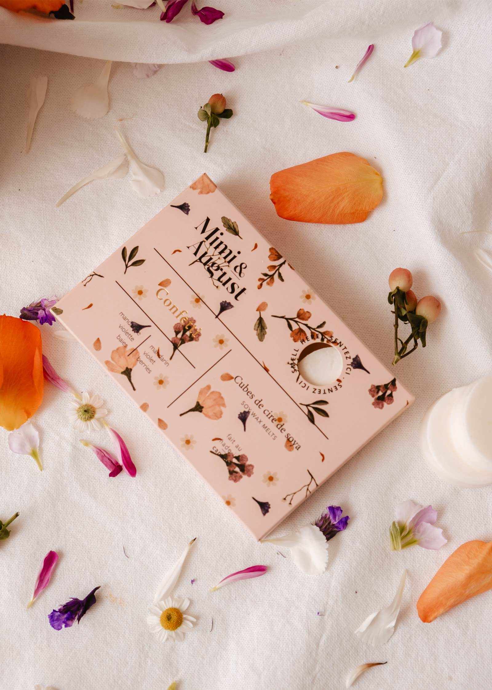 Une boîte rose à motifs floraux étiquetée « Mimi & August » repose sur un tissu blanc entourée de pétales colorés et de petites fleurs, offrant un parfum longue durée et une touche écoresponsable. Produit : Fondants de cire de soya Confetti.