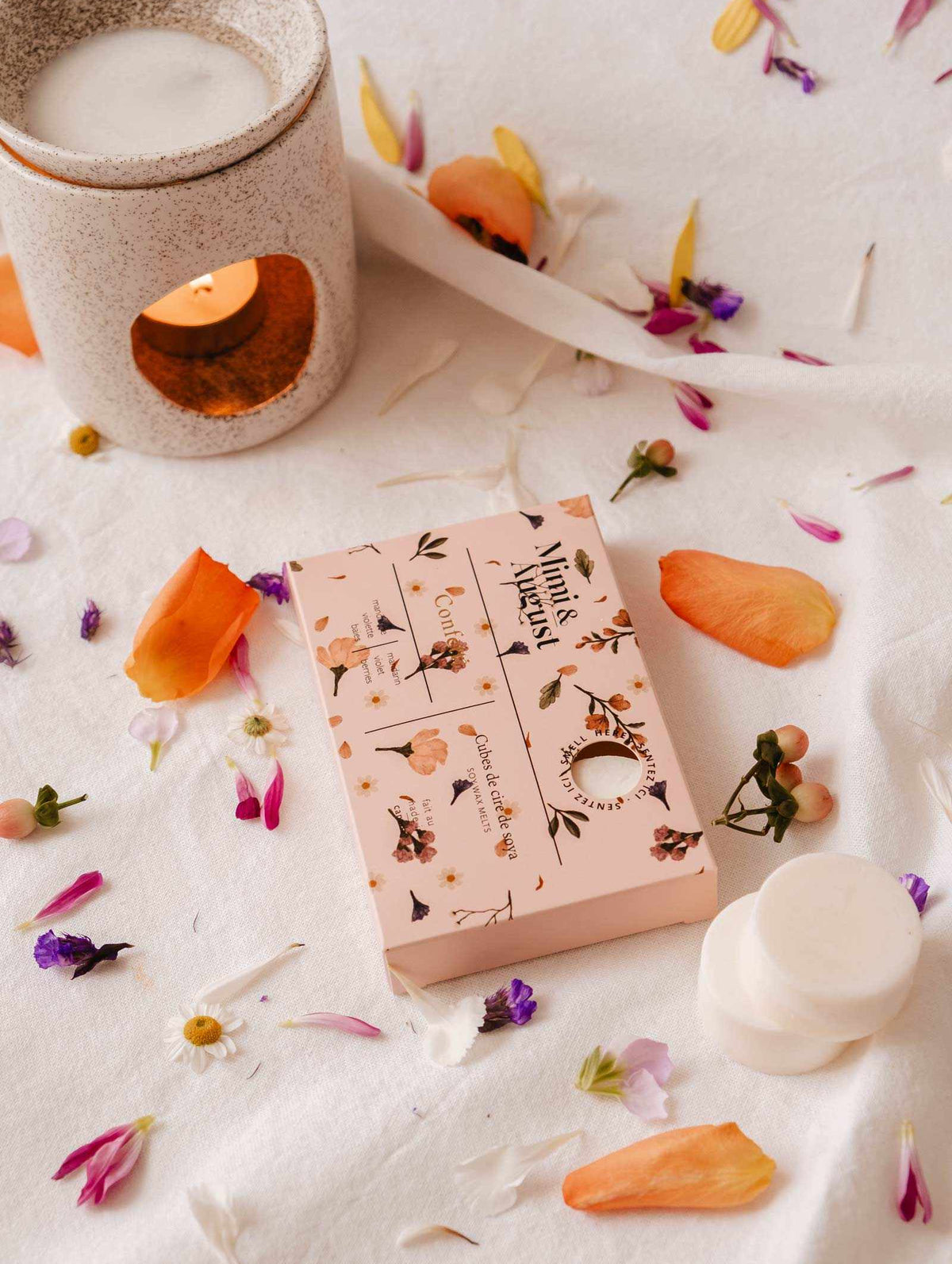 A candle warmer and a floral-patterned box rest on white fabric, surrounded by flower petals, releasing the eco-friendly, long-lasting fragrance of Mimi & August's Soy Wax Melts - Confetti.