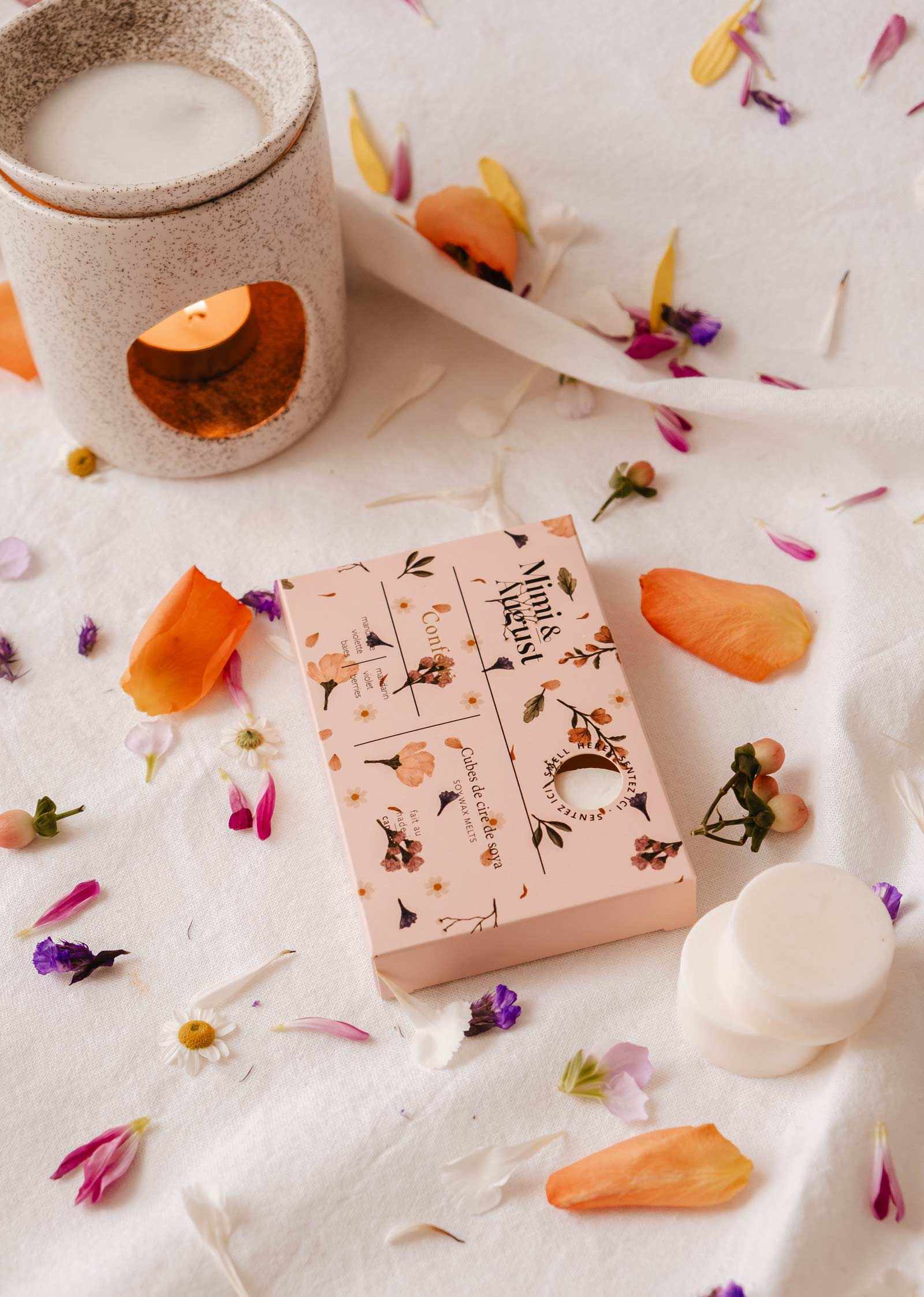 Un diffuseur à cire et une boîte à motifs floraux reposent sur un tissu blanc, entourés de pétales de fleurs, libérant le parfum écoresponsable et longue durée des fondants de cire de soya Confetti de Mimi & August.
