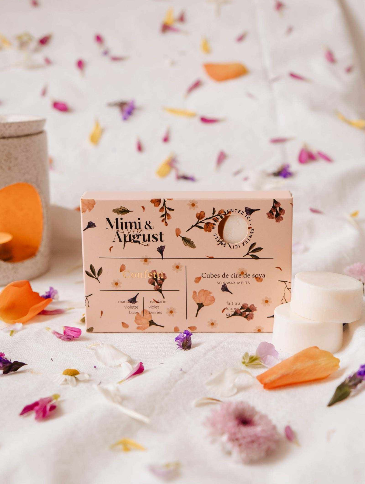 A box of Mimi & August Soy Wax Melts - Confetti sits on fabric, surrounded by colorful flower petals, creating an enchanting display with its eco-friendly composition and long-lasting fragrance.