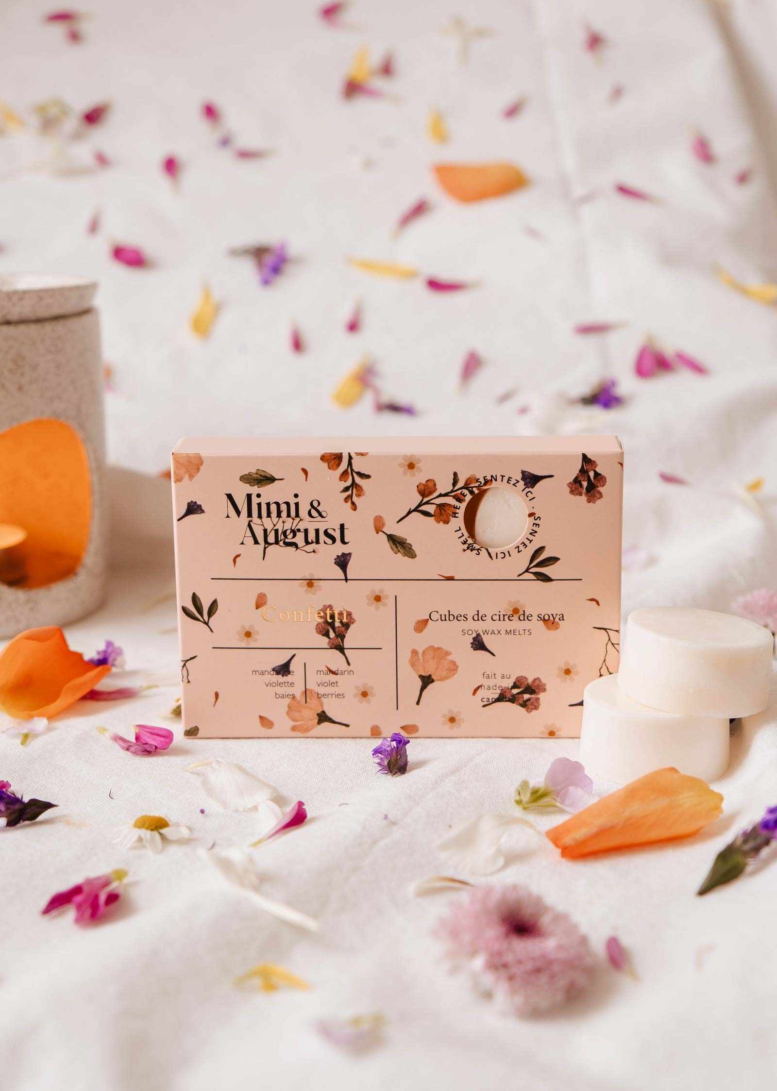 Une boîte de fondants de cire de soya Confetti de Mimi & August repose sur un tissu, entourée de pétales de fleurs colorés, créant un affichage enchanteur avec sa composition écoresponsable et son parfum longue durée.
