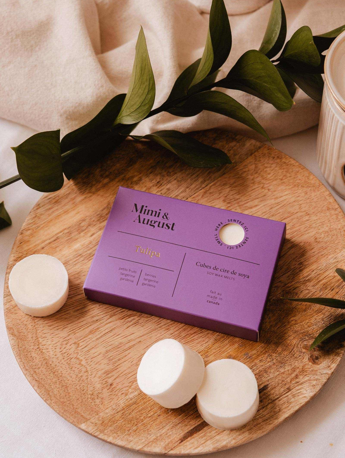 A purple Mimi & August Soy Wax Melts - Tulipa box with natural soy wax melts is shown on a round wooden tray, surrounded by greenery and loose wax melt discs.