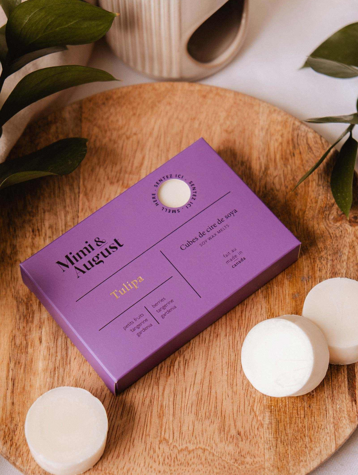 A box of Mimi & August Soy Wax Melts - Tulipa in purple rests on a round wooden tray with three white natural soy wax melts and green leaves nearby.