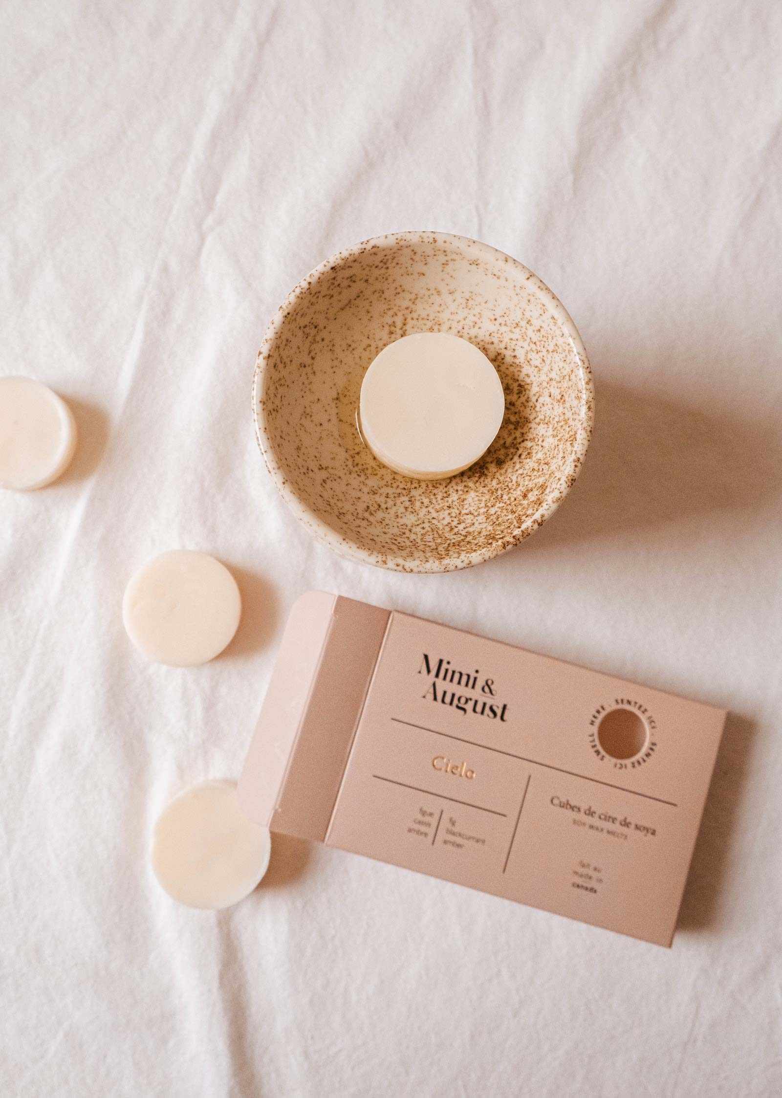 A speckled bowl cradles a cream-colored wax melt made from 100% soy. An open "fig wax melts - Cielo" package by Mimi & August promises lasting fragrance, with extra melts laid on a white fabric.