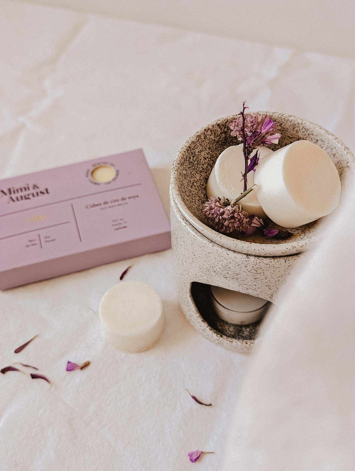 A beige wax melt burner with eco-friendly Mimi & August Soy Wax Melts - Lilac and dried flowers sits by a matching lilac box, filling the air with long-lasting fragrance atop a white fabric surface.