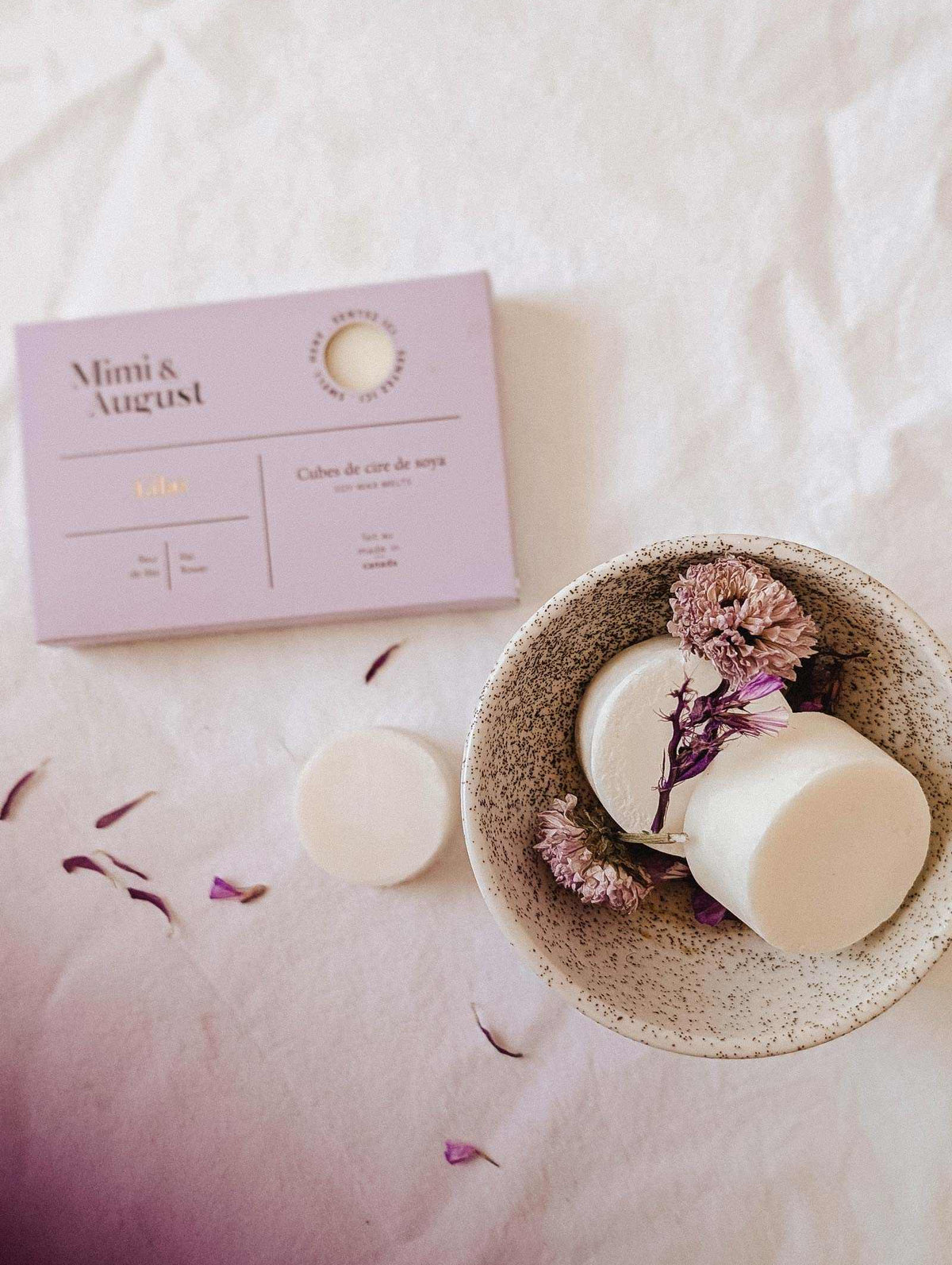 A speckled bowl with two Mimi & August Soy Wax Melts - Lilac and dried flowers sits beside a lavender box on a white surface, providing an eco-friendly, long-lasting fragrance for your space.