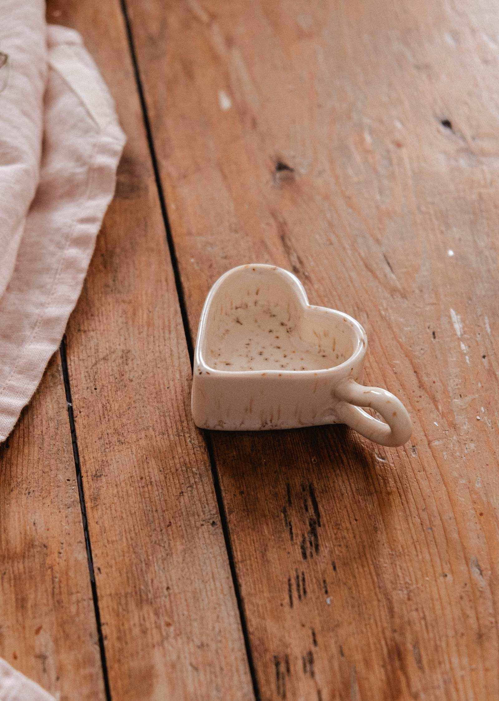 The Speckled Heart Tealight Holder by Mimi & August sits on a rustic wooden surface beside a folded beige cloth.