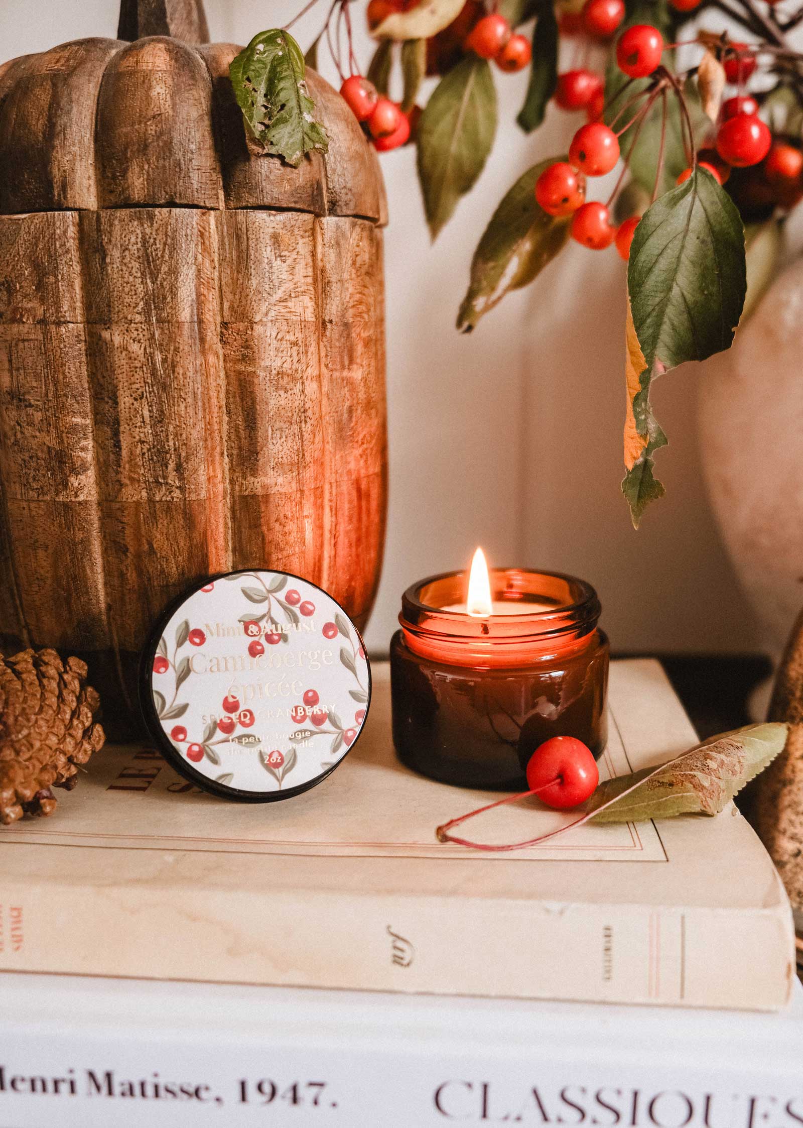 The Mimi & August Spiced Cranberry - 2 oz candle, in a berry-themed tin, is arranged with pinecones and red berries on stacked books for a cozy, festive display perfect for autumn or the holidays.