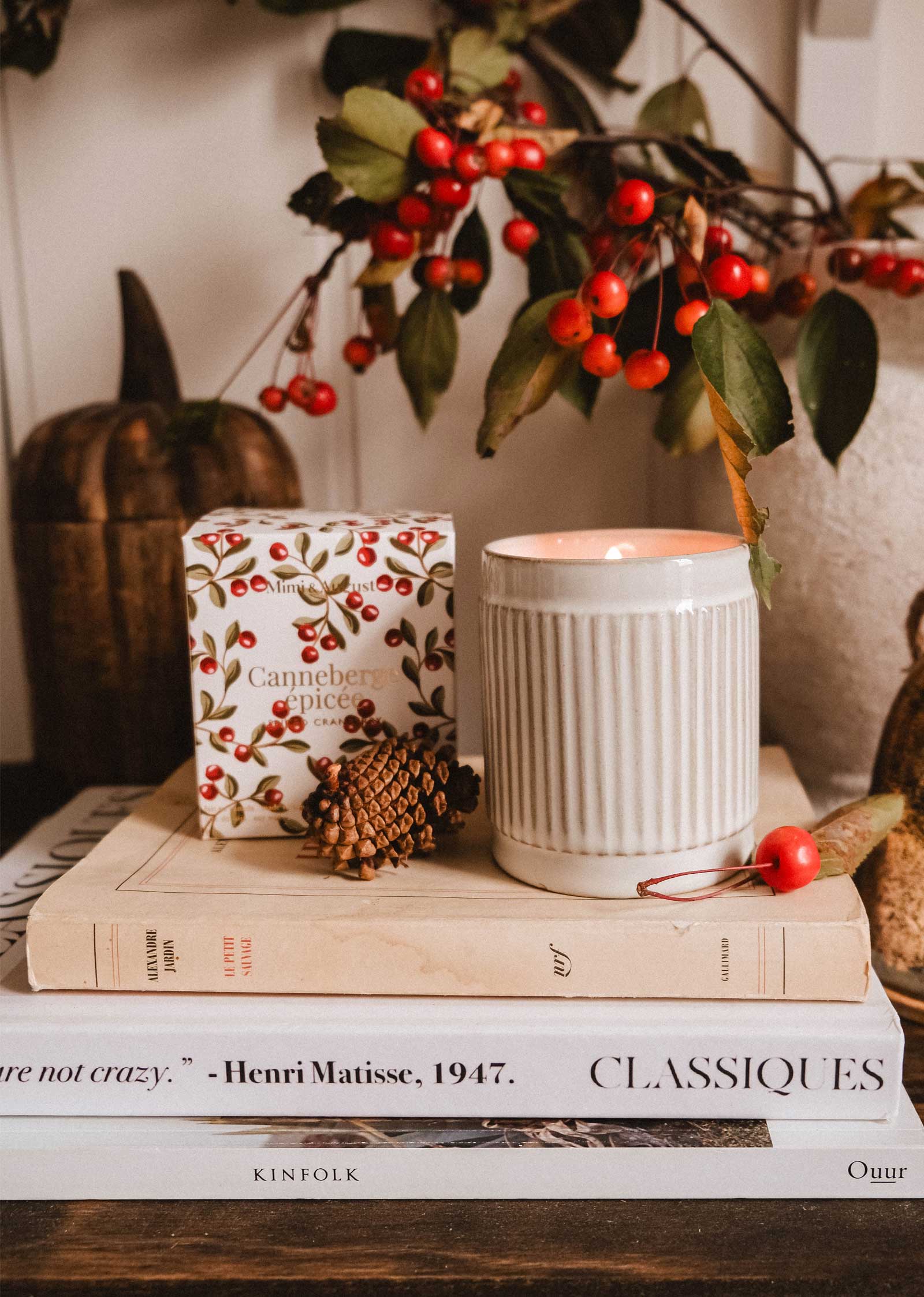 The Mimi & August Spiced Cranberry - Christmas Candle glows beside stacked books, a pinecone, and red berries with leaves, adding festive warmth to the scene.