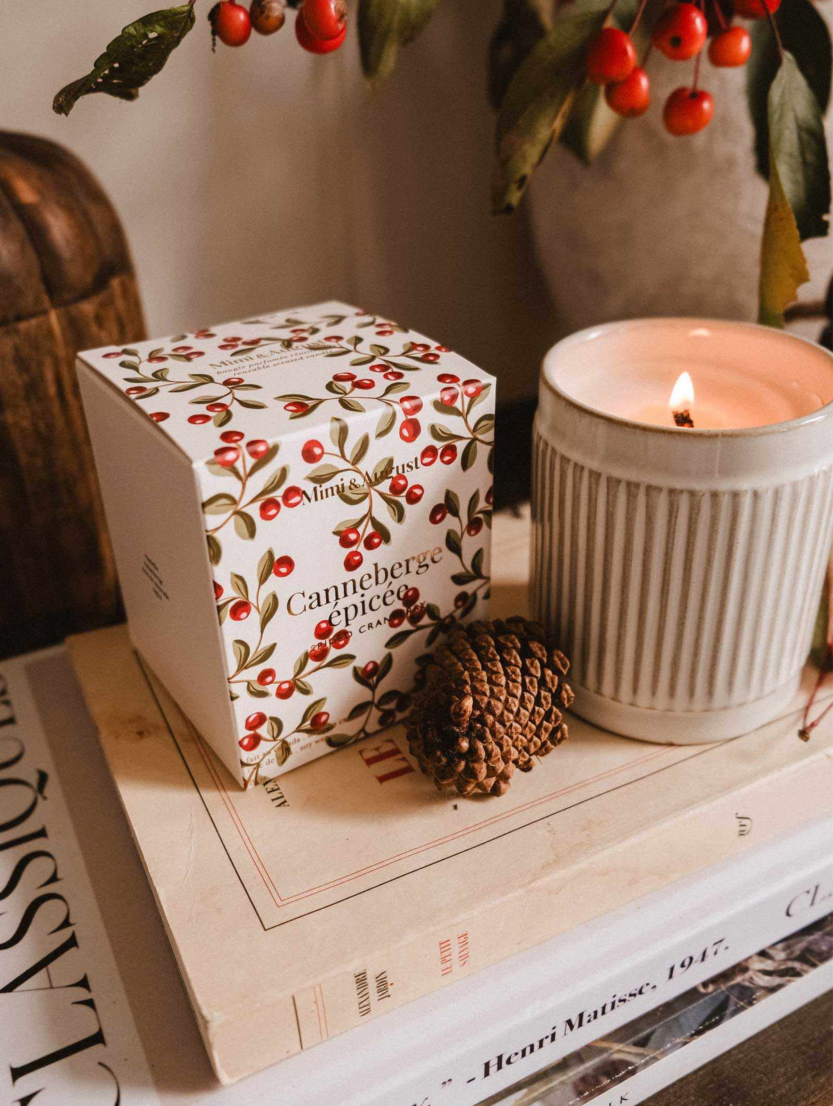 A lit Mimi & August Spiced Cranberry - Christmas Candle, a pinecone, and a cranberry-themed box rest on two stacked art books beside a branch with red berries.