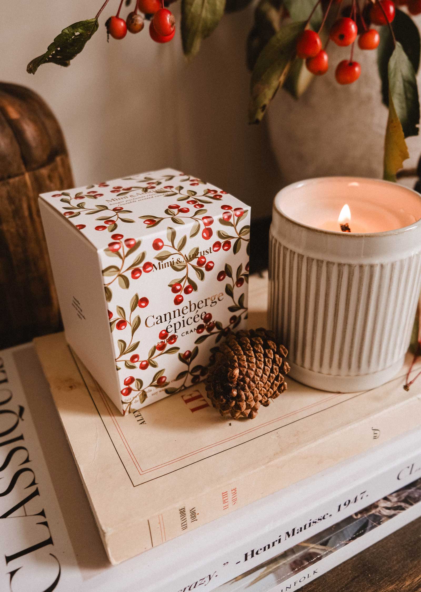 A lit Mimi & August Spiced Cranberry - Christmas Candle, a pinecone, and a cranberry-themed box rest on two stacked art books beside a branch with red berries.