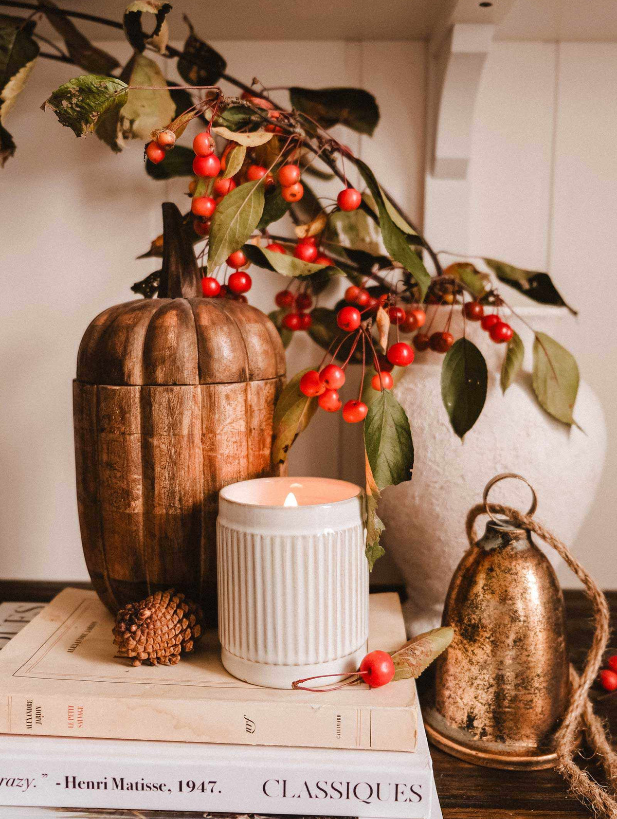 A wooden pumpkin, a gold bell, books, and a Mimi & August Spiced Cranberry - Christmas Candle in a white jar sit on a table next to a white vase filled with red-berried branches.