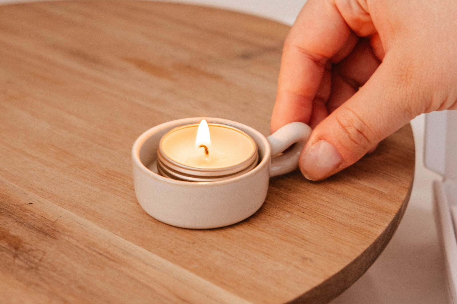 A hand places a lit tealight in the Mimi & August Tealight Holder, featuring a white ceramic base on a round wooden surface—an ideal accent for rustic decor.