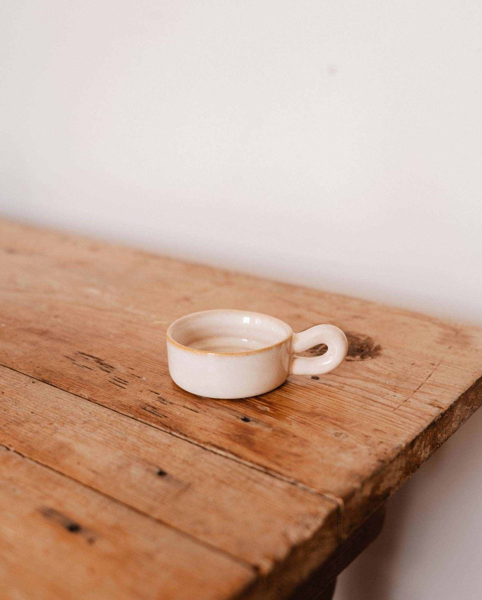 The Mimi & August Tealight Holder, a small empty beige ceramic piece with a handle, rests on a rustic wooden table against a plain white wall.