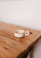 The Mimi & August Tealight Holder, a small empty beige ceramic piece with a handle, rests on a rustic wooden table against a plain white wall.