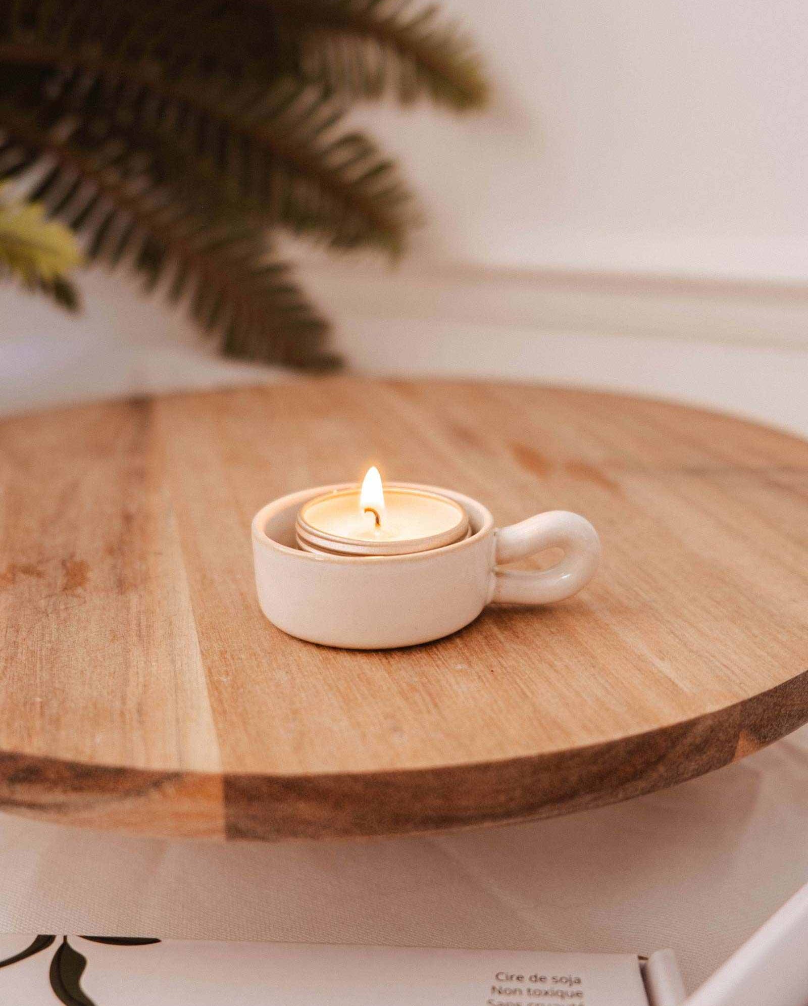 A lit tealight sits in the Mimi & August Tealight Holder, a small white ceramic cup on a wooden surface, bringing charm to rustic decor. A leafy plant adds a natural touch in the background.