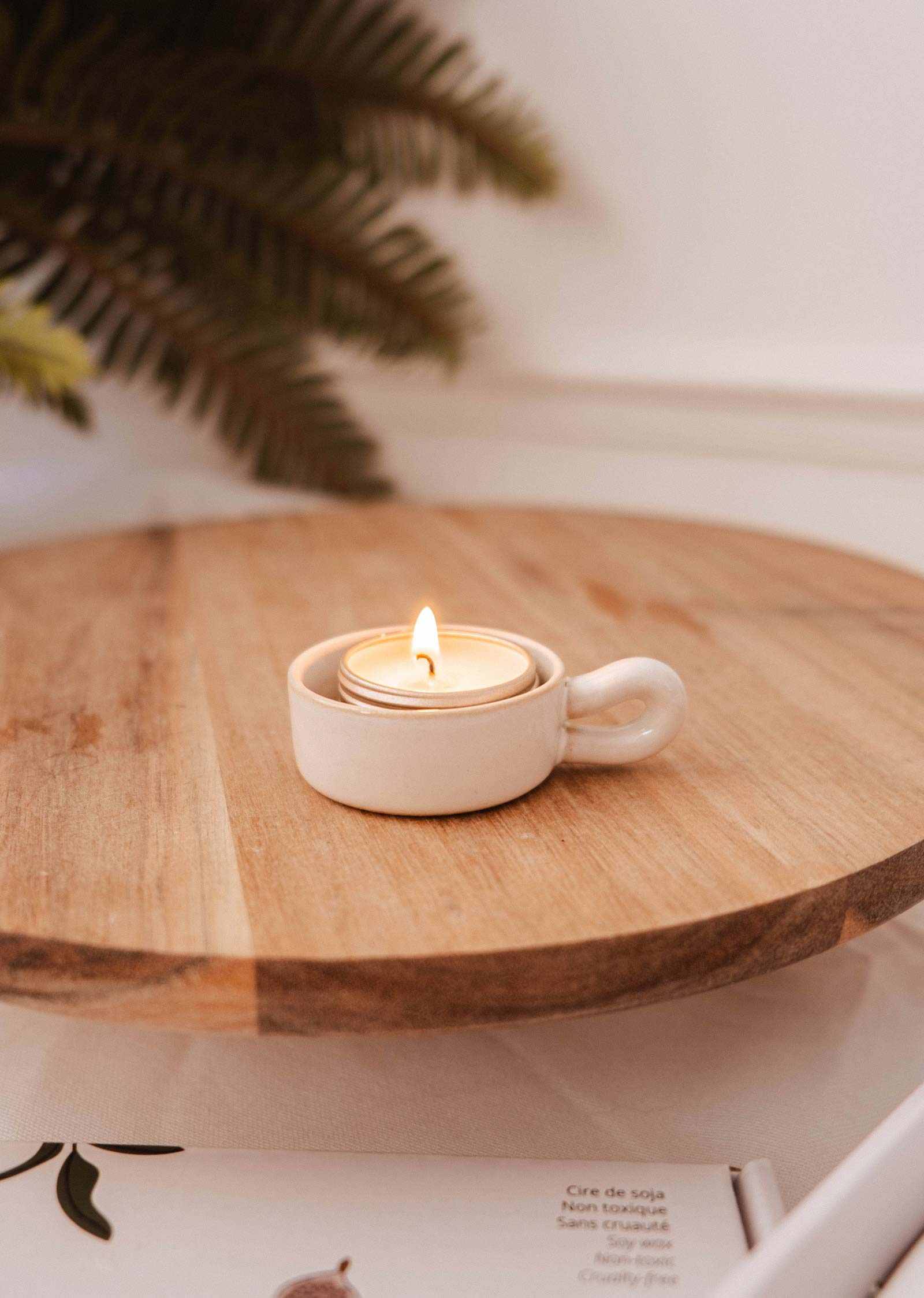 A lit tealight sits in the Mimi & August Tealight Holder, a small white ceramic cup on a wooden surface, bringing charm to rustic decor. A leafy plant adds a natural touch in the background.