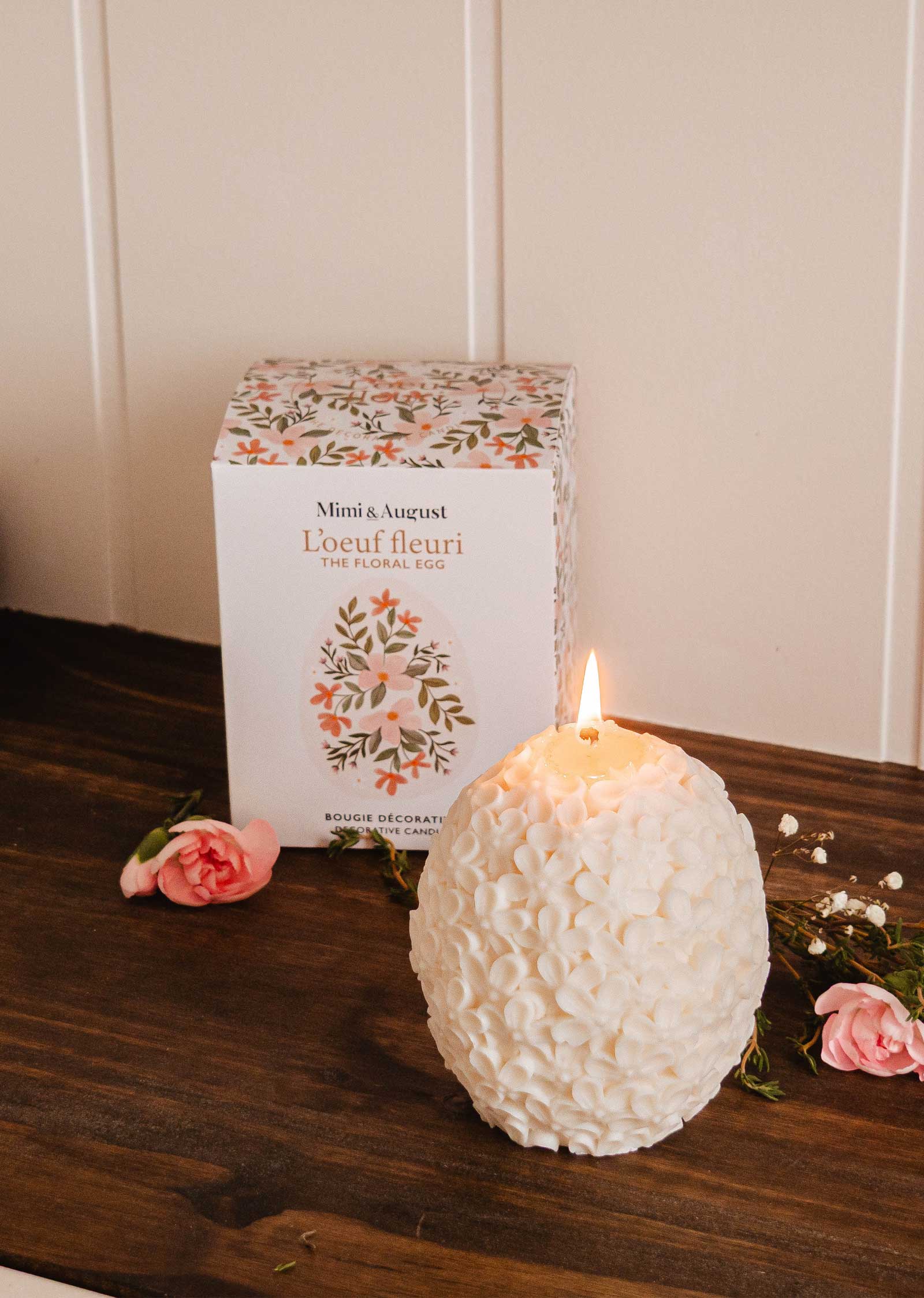 The Floral Egg - Decorative Candle by Mimi & August boasts ornate floral patterns on a white egg-shaped design. Display it lit on a wooden table with its matching floral box and scattered pink flowers for an elegant décor touch.