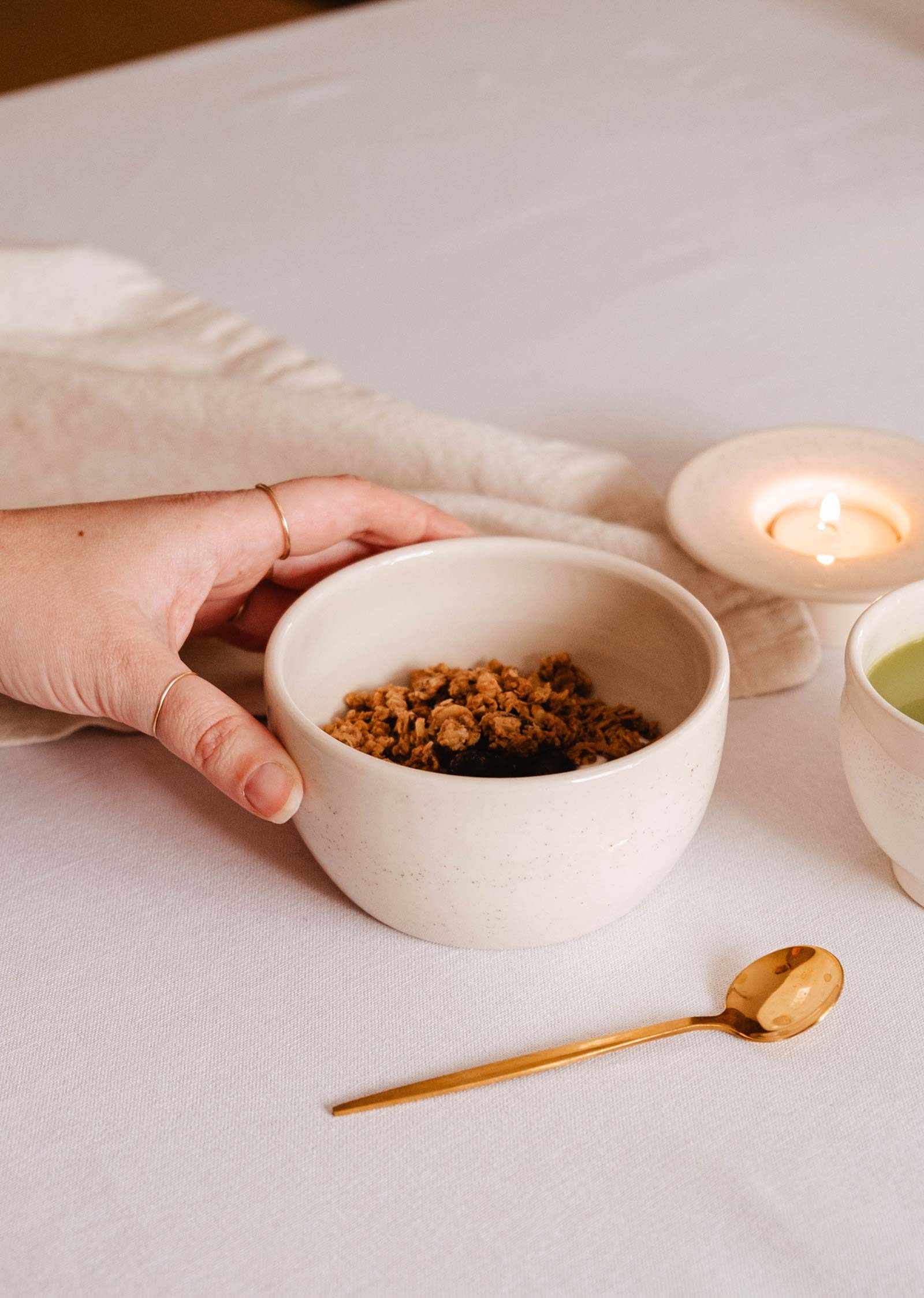 Une main tient Le bol du matin – céramique artisanale de Mimi & August, rempli de granola, posé à côté d'une cuillère dorée, d'une tasse et d'un lampion allumé sur une nappe blanche.
