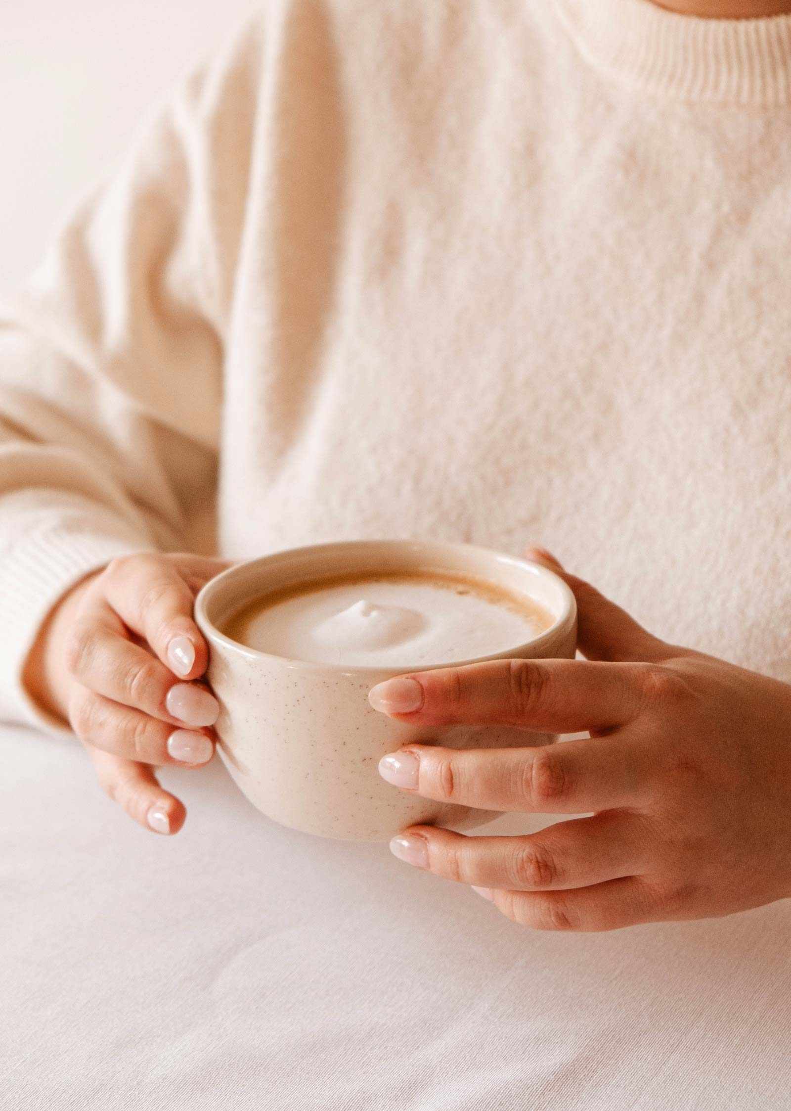 Une personne en chandail crème tient un cappuccino mousseux dans Le bol du matin – céramique artisanale de Mimi & August, à la finition crème mouchetée, posé sur une table blanche.