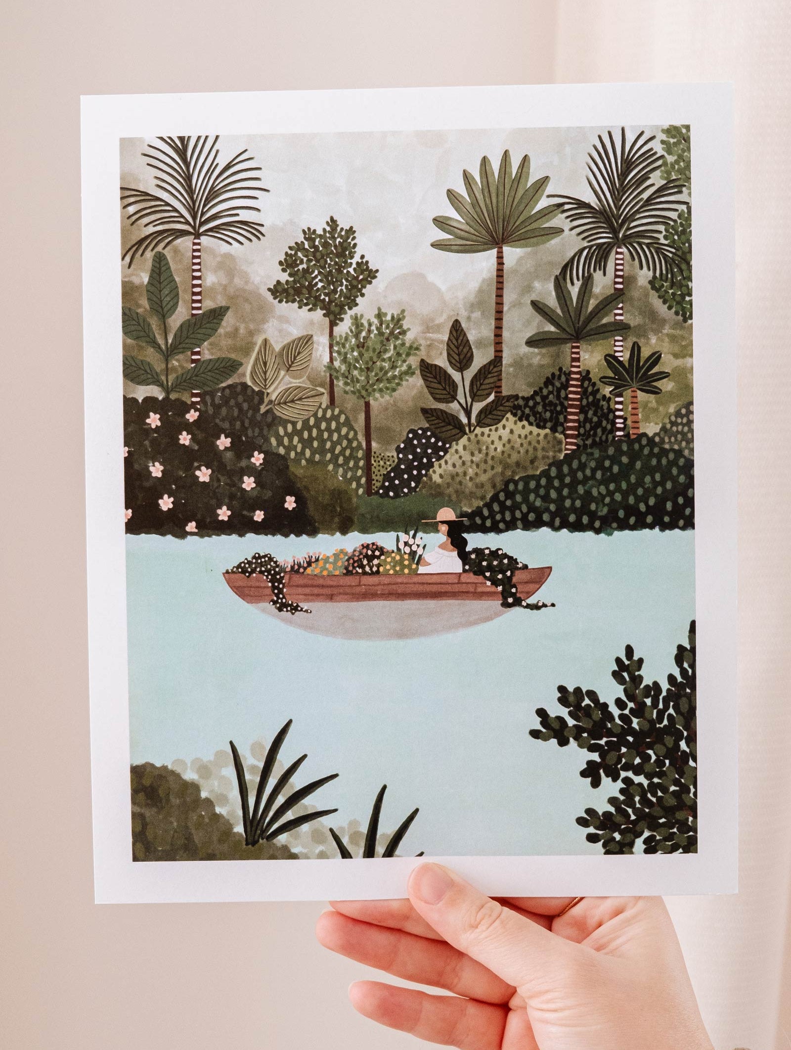 An Mimi & August art print capturing the natural tranquility of a scenic journey, with a hand holding a picture of The Lake Art Print among trees.