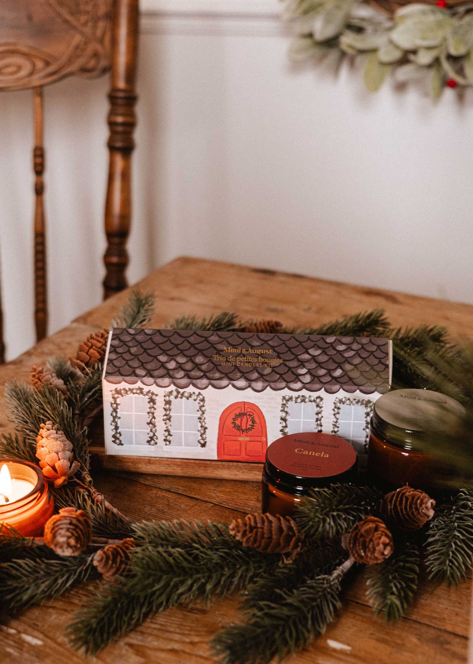 The Little Christmas Box by Mimi & August sits on a wooden table with pine branches, pine cones, a lit holiday-scented candle, and two small jars.