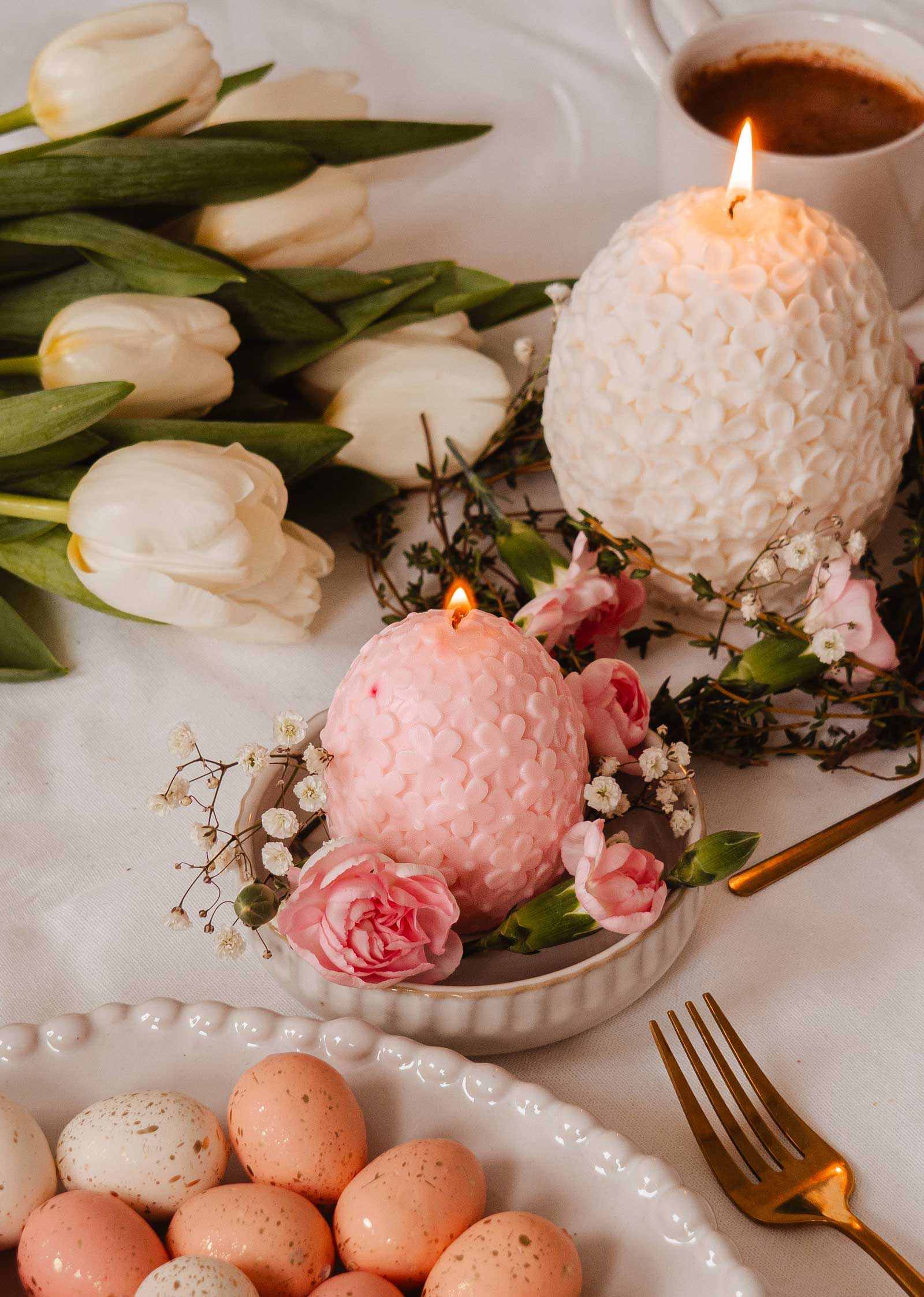 Mimi & August's The Little Floral Egg - Decorative Candle in blush pink adds charm to the table with white tulips, coffee, a gold fork, and speckled eggs, releasing a delightful floral scent into the air.