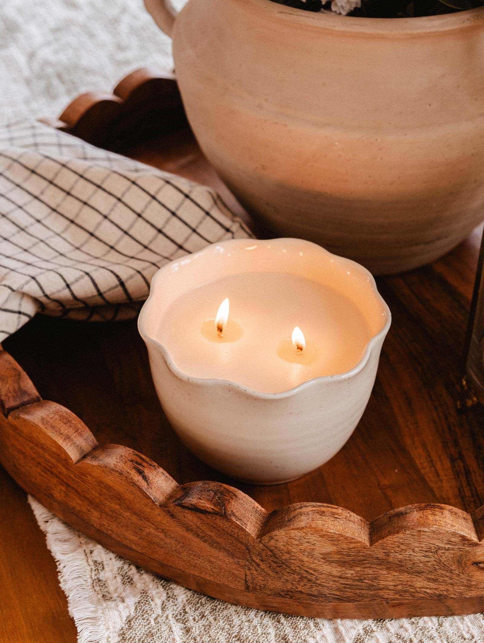 The Petal Bowl Candle by Mimi & August features two lit wicks in a handcrafted ceramic bowl, set on a wooden tray beside a checkered cloth and beige pot—an ideal Mother's Day gift.