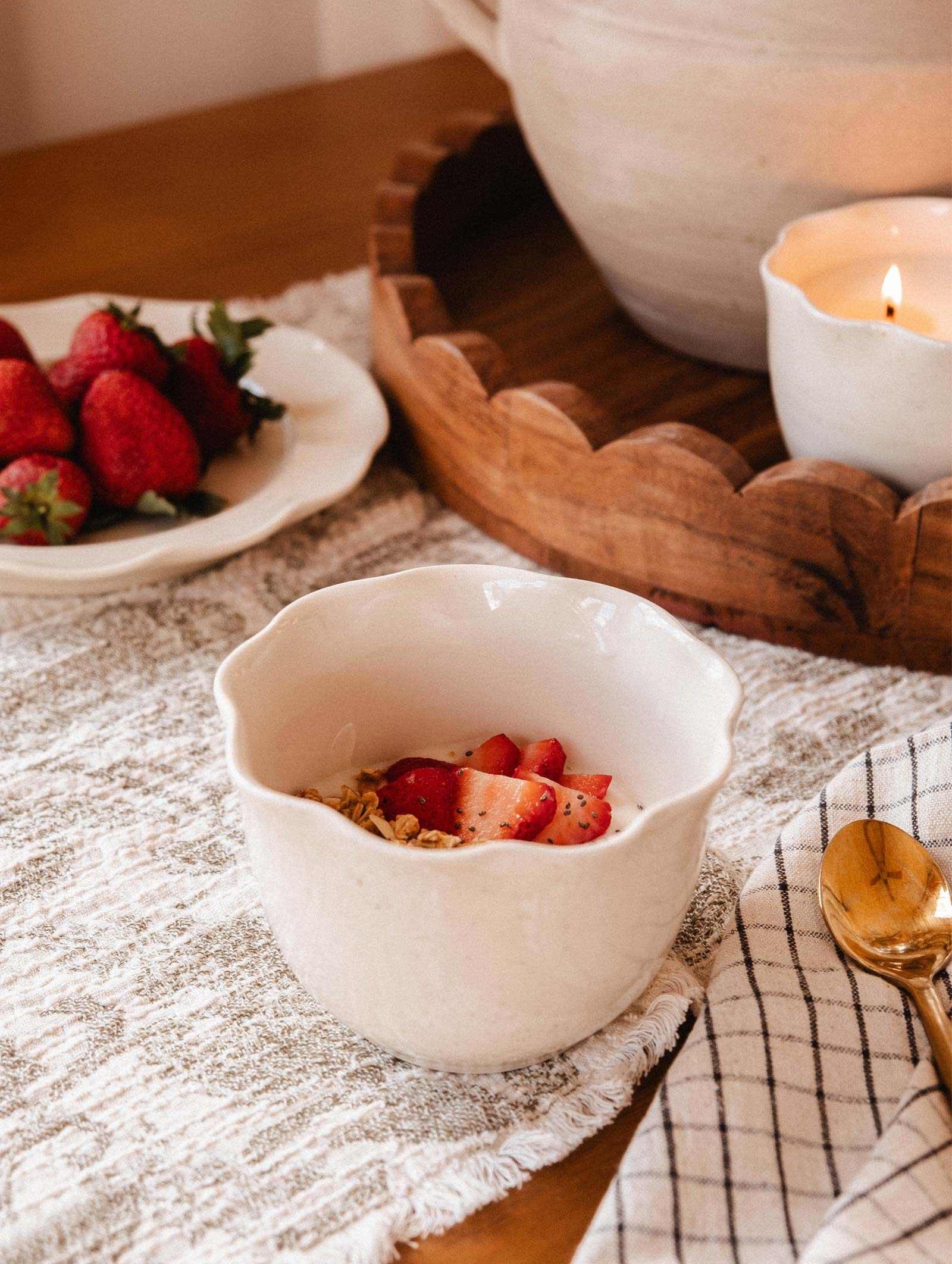 A white bowl with yogurt, granola, and strawberries rests on a textured cloth beside a gold spoon and checkered napkin; in the background, fresh strawberries and Mimi & August's The Petal Bowl Candle add a soft glow.