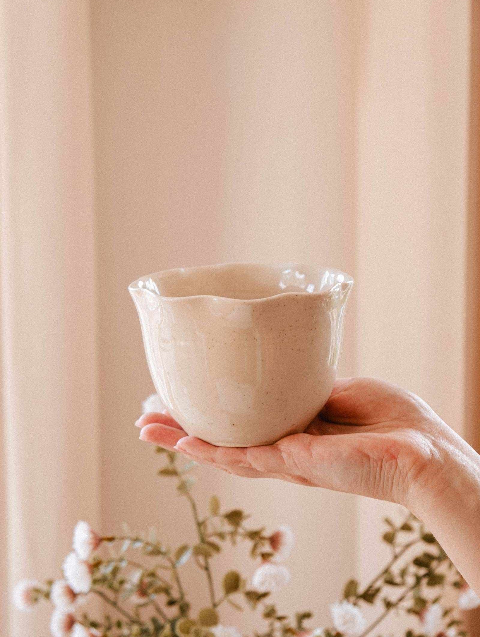 A hand holds The Petal Bowl Candle by Mimi & August, a light beige, scalloped-edge handcrafted ceramic candle with a glossy finish—ideal as a limited edition piece or thoughtful Mother's Day gift; blurred flowers in the background.