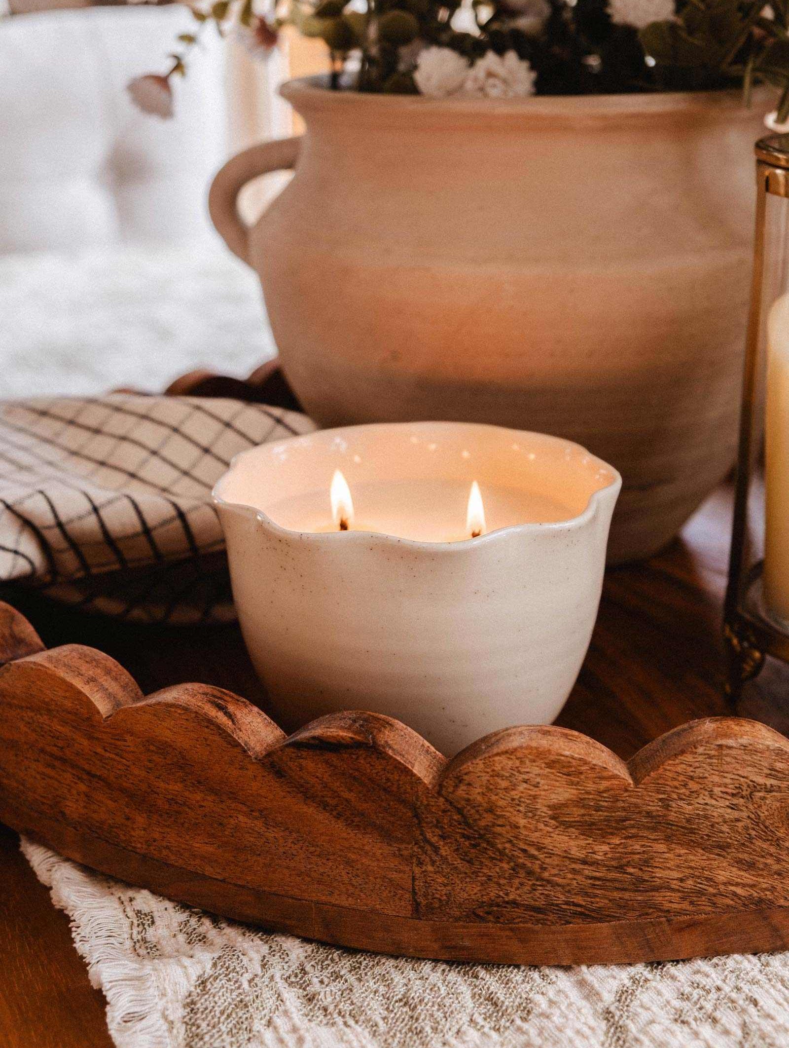 The Petal Bowl Candle by Mimi & August features two flames in a white ceramic holder. Set on a lace tablecloth beside a wooden tray, checked napkin, and beige floral vase, it makes a perfect Mother's Day gift.