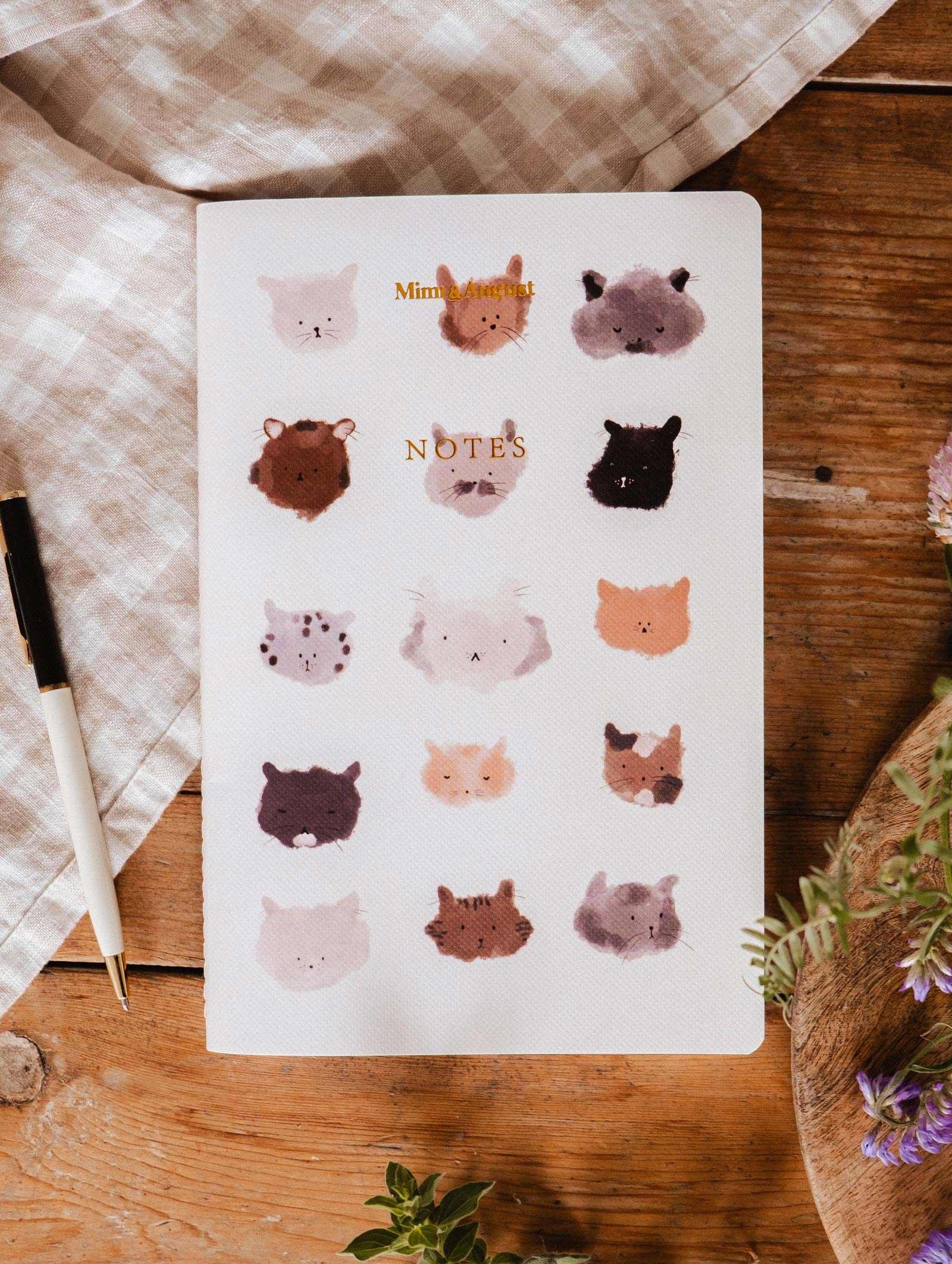 The Purrfect - Notebook by Mimi & August features illustrated cat faces on the cover and is shown on a wooden table next to a pen, checkered cloth, and flowers.
