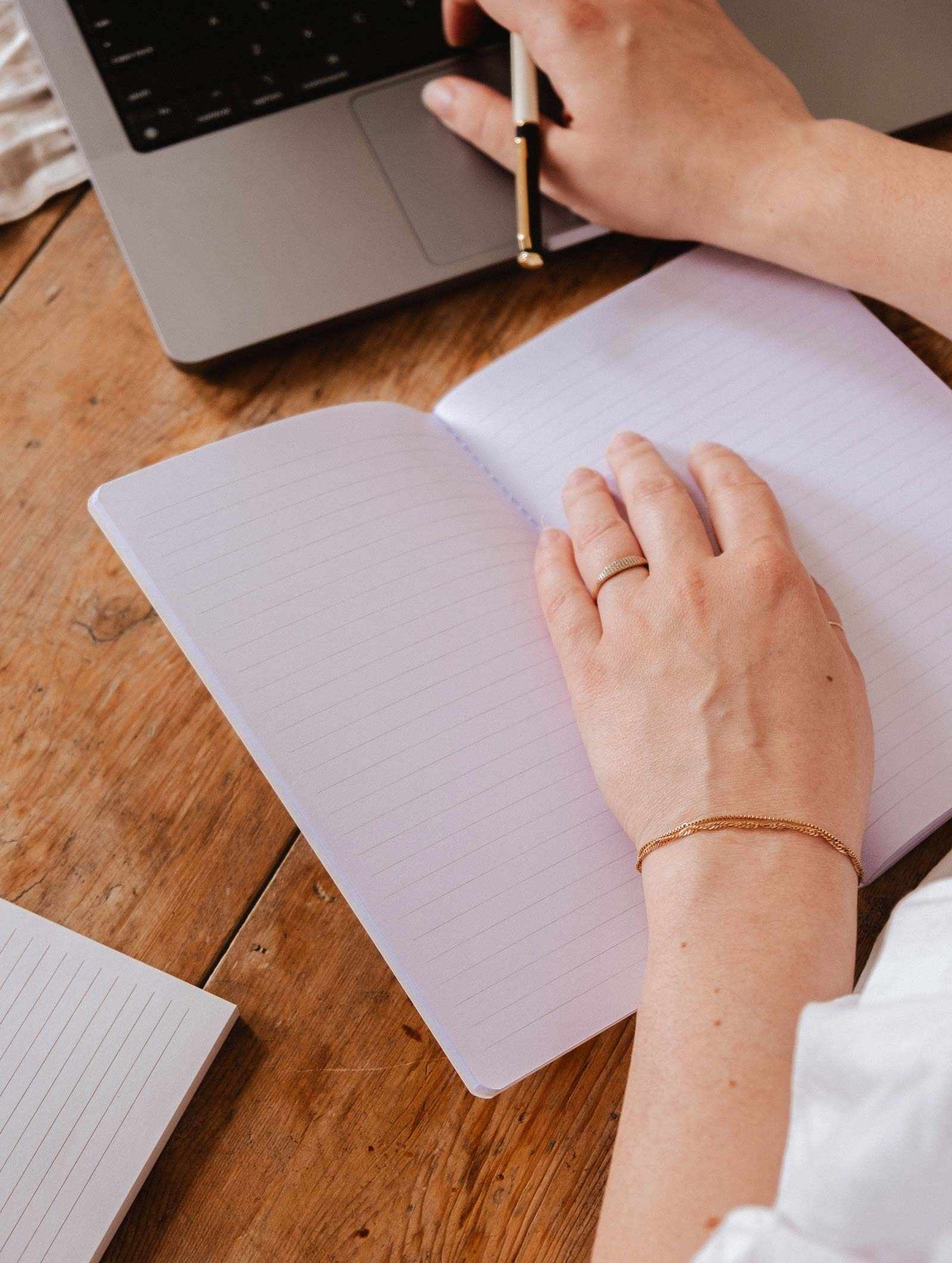 Someone writes in The Purrfect - Notebook by Mimi & August, with cute stationery next to an open laptop on a wooden desk.