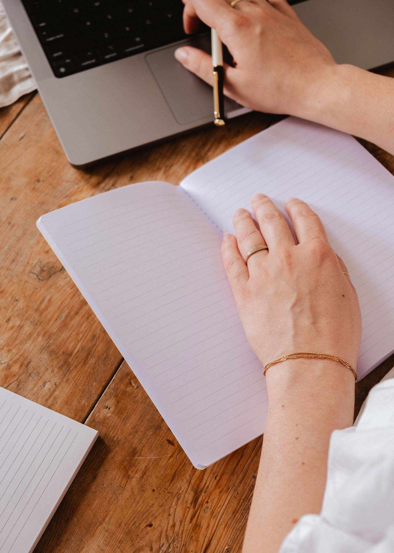 Someone writes in The Purrfect - Notebook by Mimi & August, with cute stationery next to an open laptop on a wooden desk.