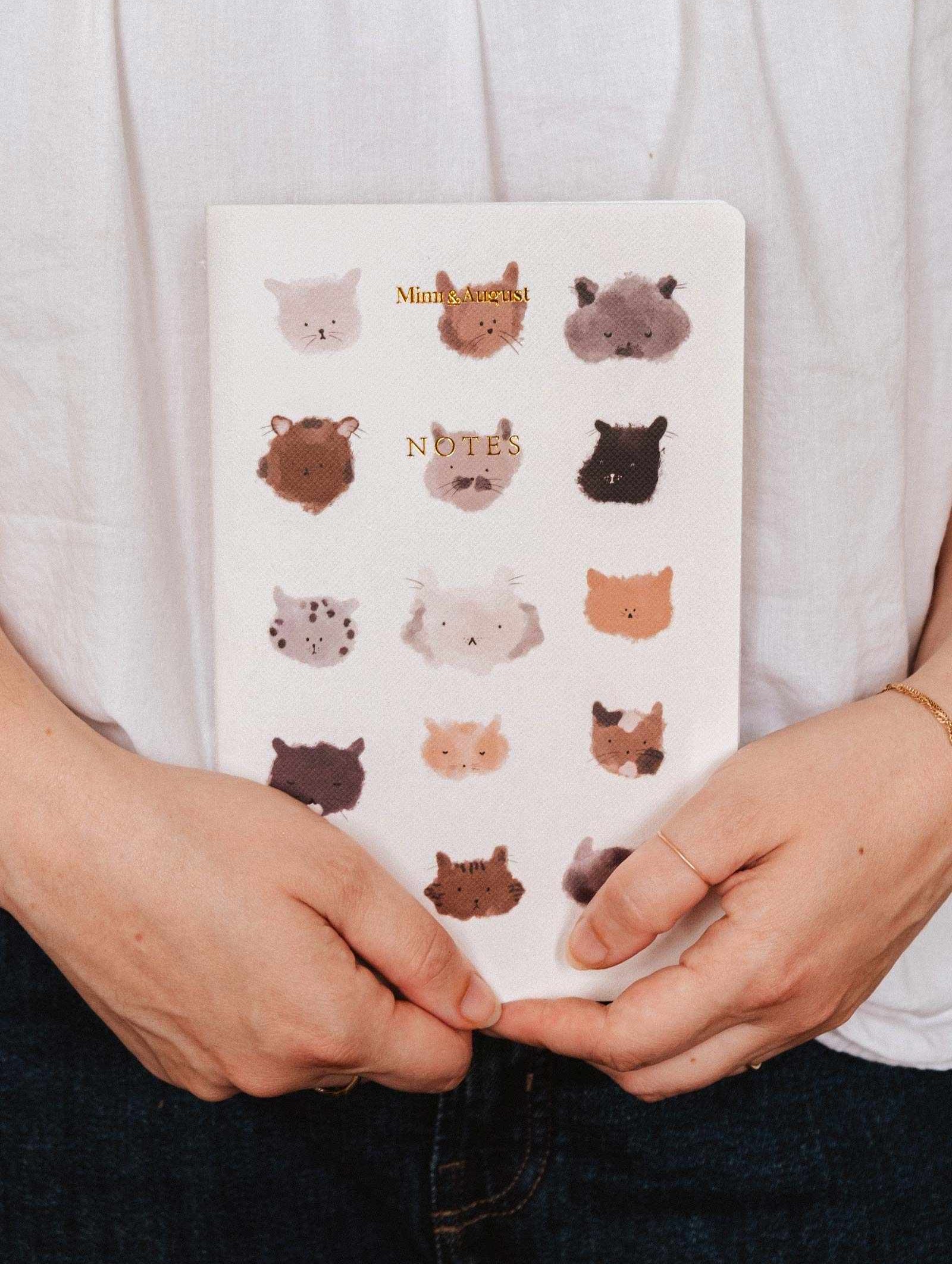Someone holds The Purrfect - Notebook by Mimi & August, a white notebook with illustrated animal faces in a grid on the cover—ideal for cute stationery lovers.