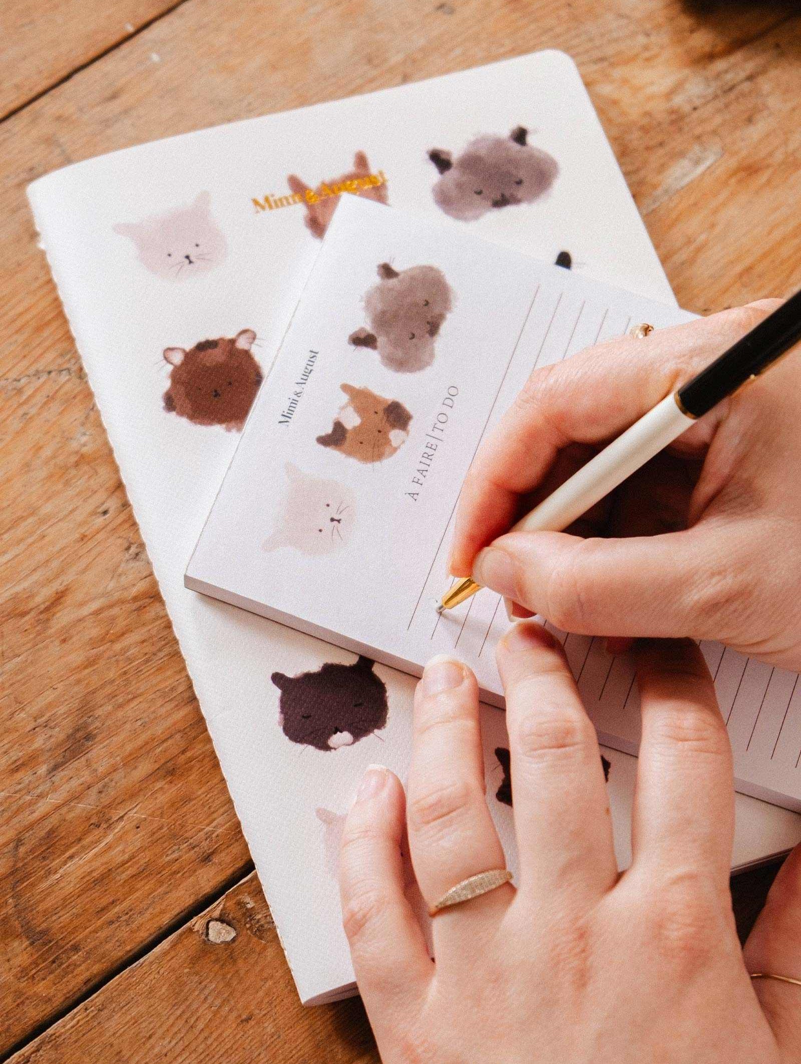Someone writes on the Mimi & August Purrfect Notepad, featuring adorable cat illustrations, with a matching notebook underneath on a wooden table.