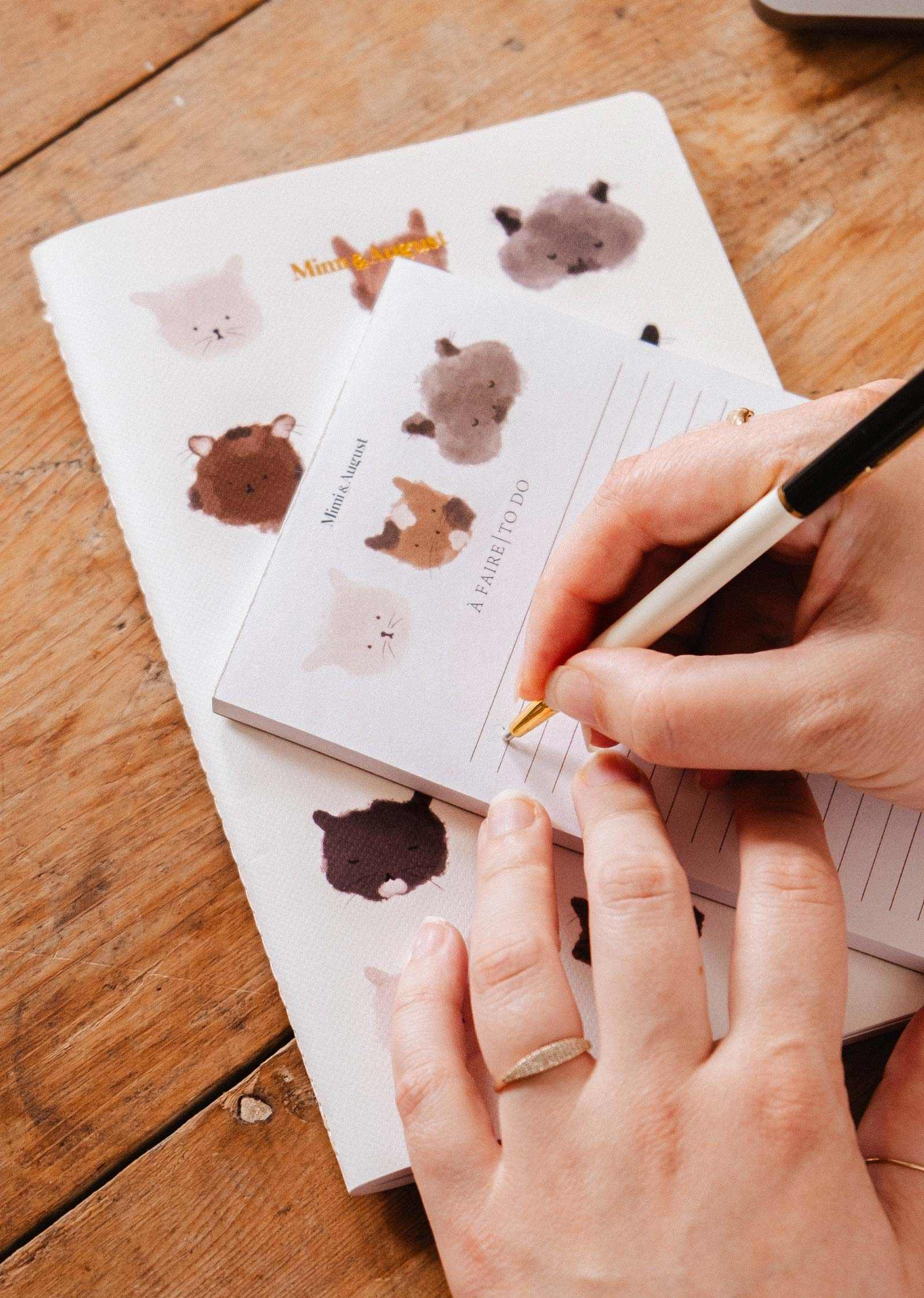 Someone writes on the Mimi & August Purrfect Notepad, featuring adorable cat illustrations, with a matching notebook underneath on a wooden table.