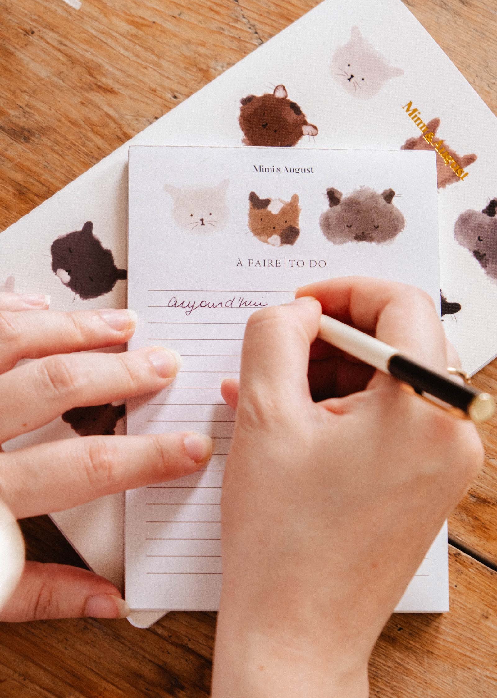 Someone writes on the Mimi & August Purrfect - Notepad, adorned with illustrated kitty faces, using a black and gold pen on a wooden surface.