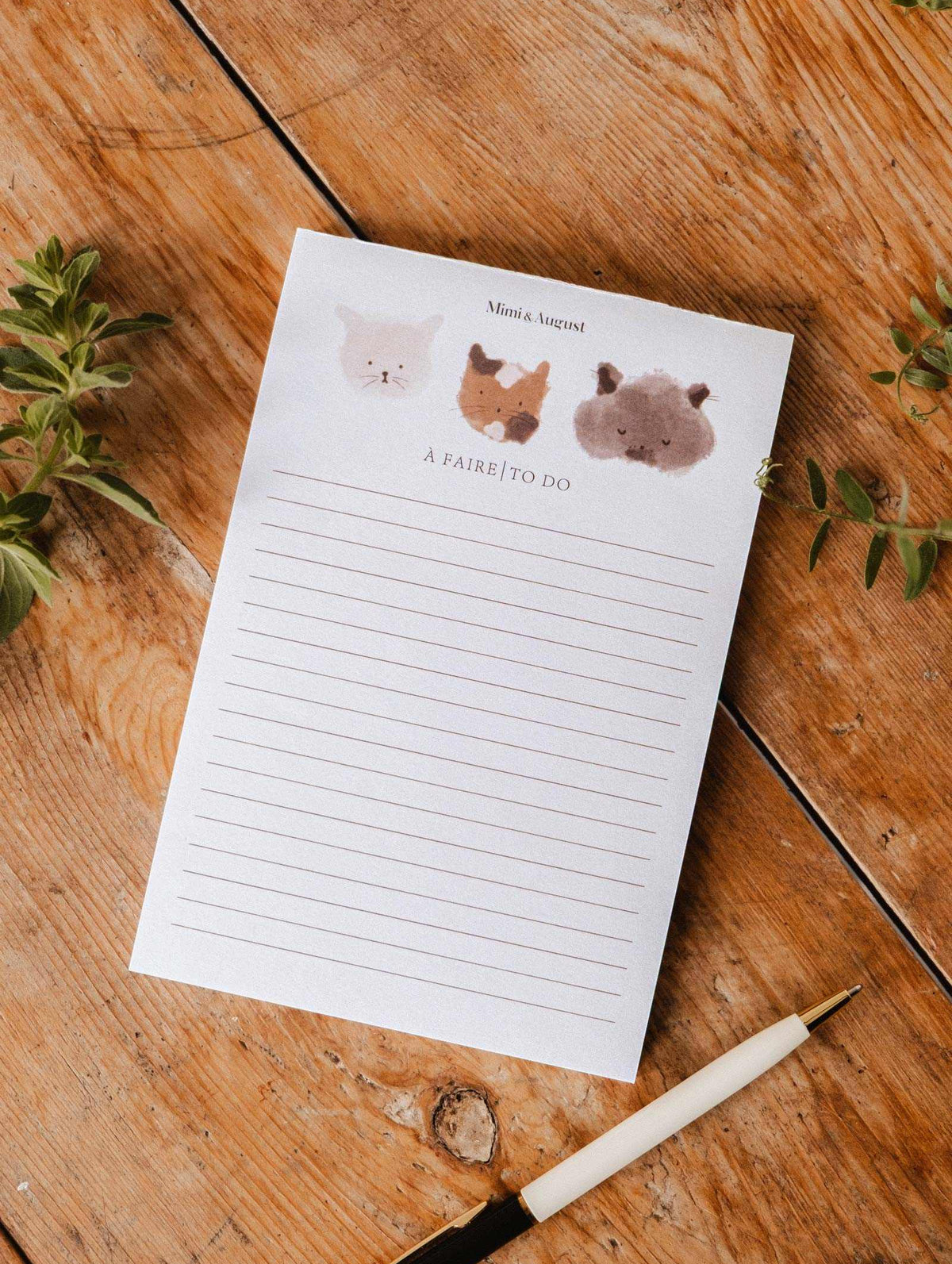 The "Purrfect - Notepad" by Mimi & August features adorable kitty illustrations at the top and lined pages, perfect for jotting down to-dos. Shown on a wooden surface beside a pen and green plants.
