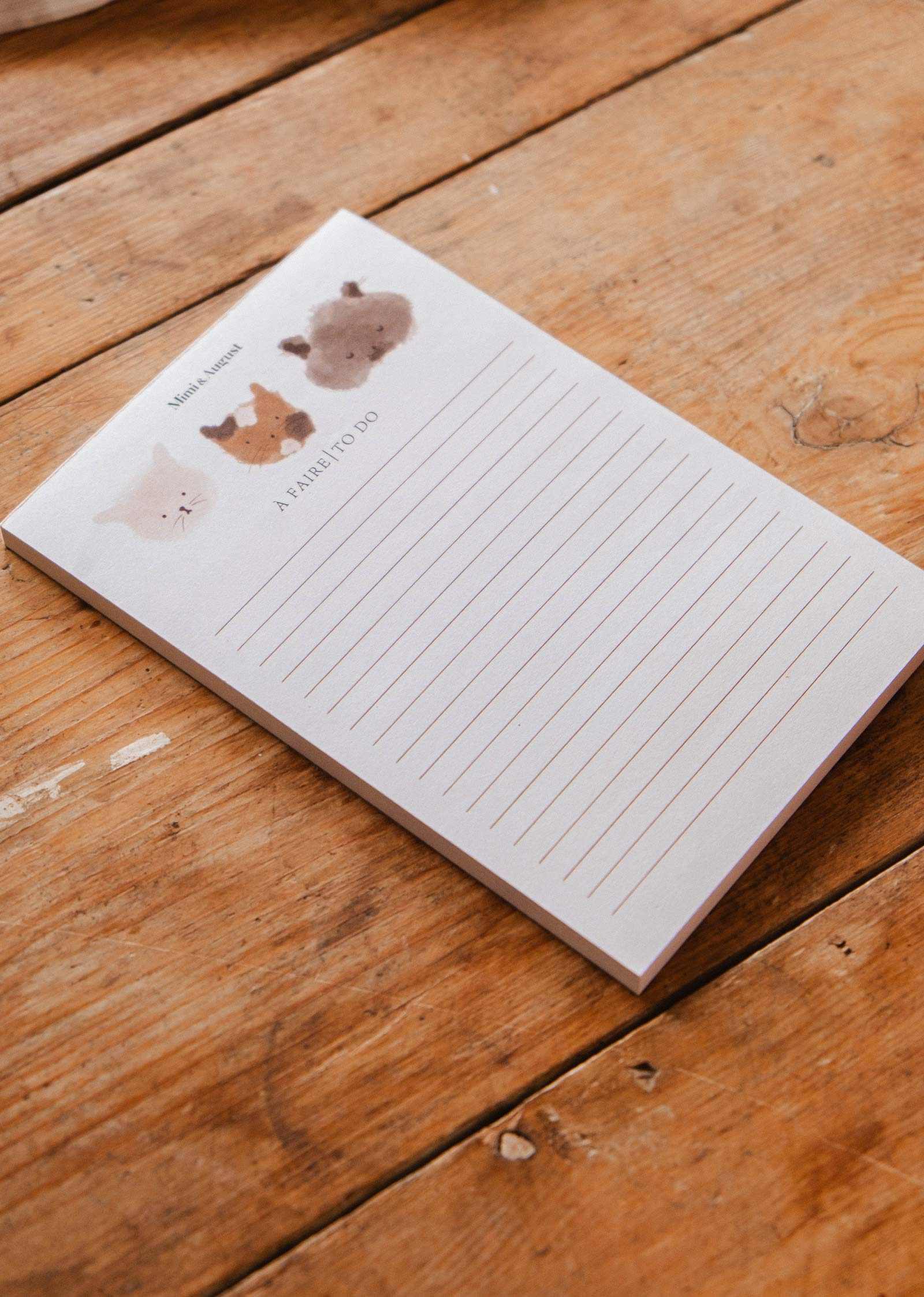 The Purrfect - Notepad by Mimi & August features lined pages with cartoon animal faces, including a kitty illustration at the top, making it a cute stationery item for your desk.