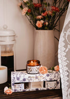 A lit floral candle in an amber jar is on a tray with a coffee cup, pink flowers in a vase, and the elegant packaging of Mimi & August's The Secret Garden Box Set.